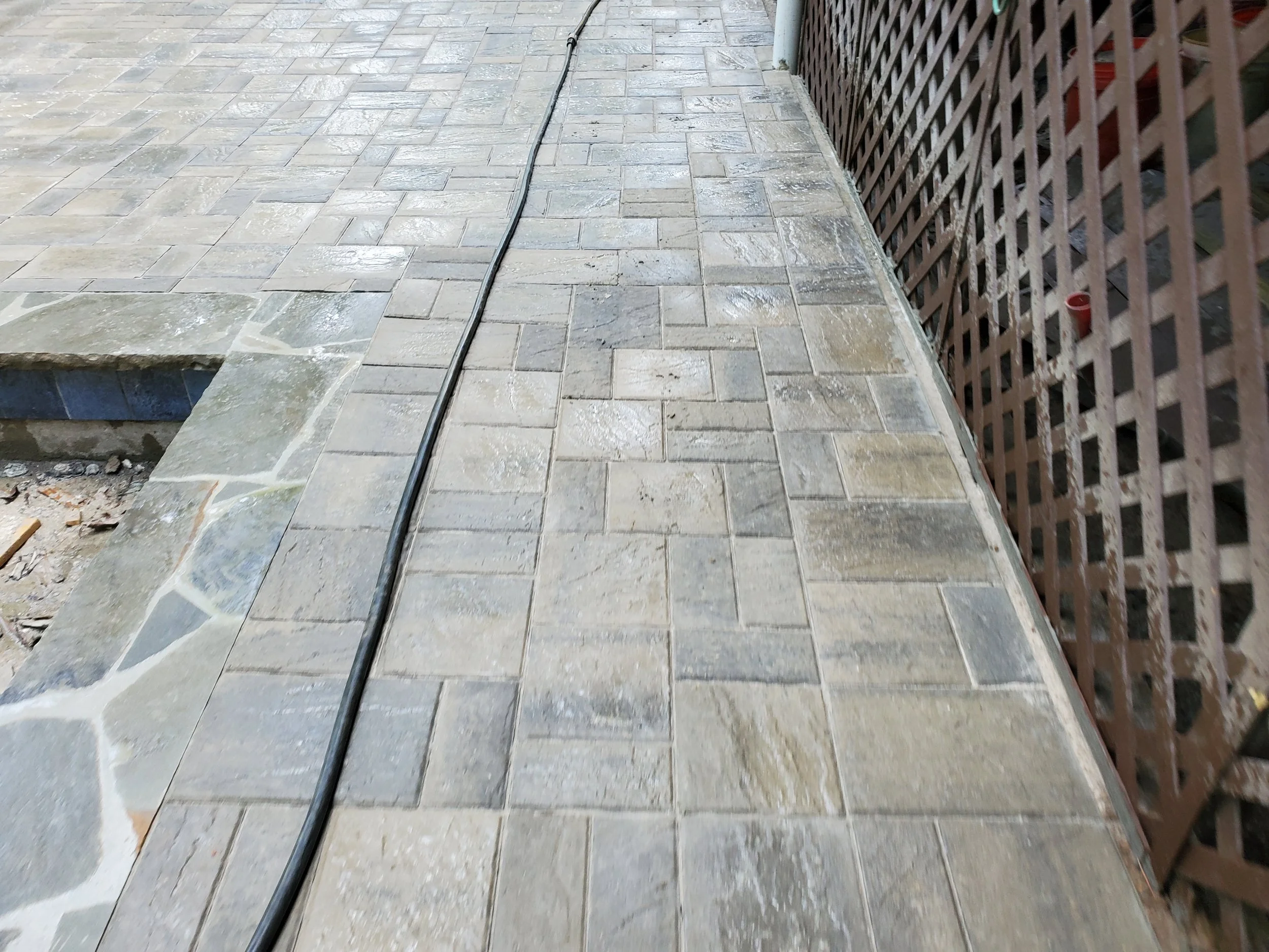 Photo of a tiled outdoor patio area with a black cable running across it and a brown wooden lattice fence on the right side.