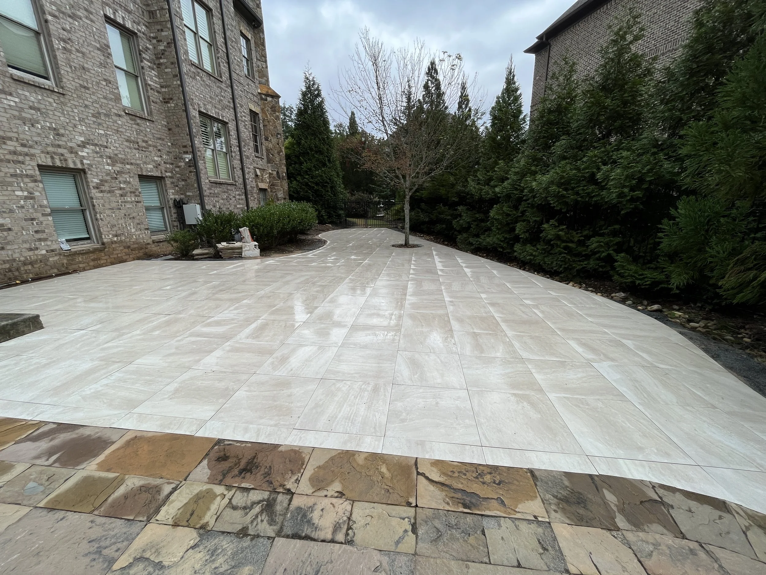 A backyard patio with square light beige tiles, bordered at the front with darker, multi-colored stone tiles. There is a leafless tree planted in a small circular bed and surrounded by mulch. To the left, there are some construction materials near a 