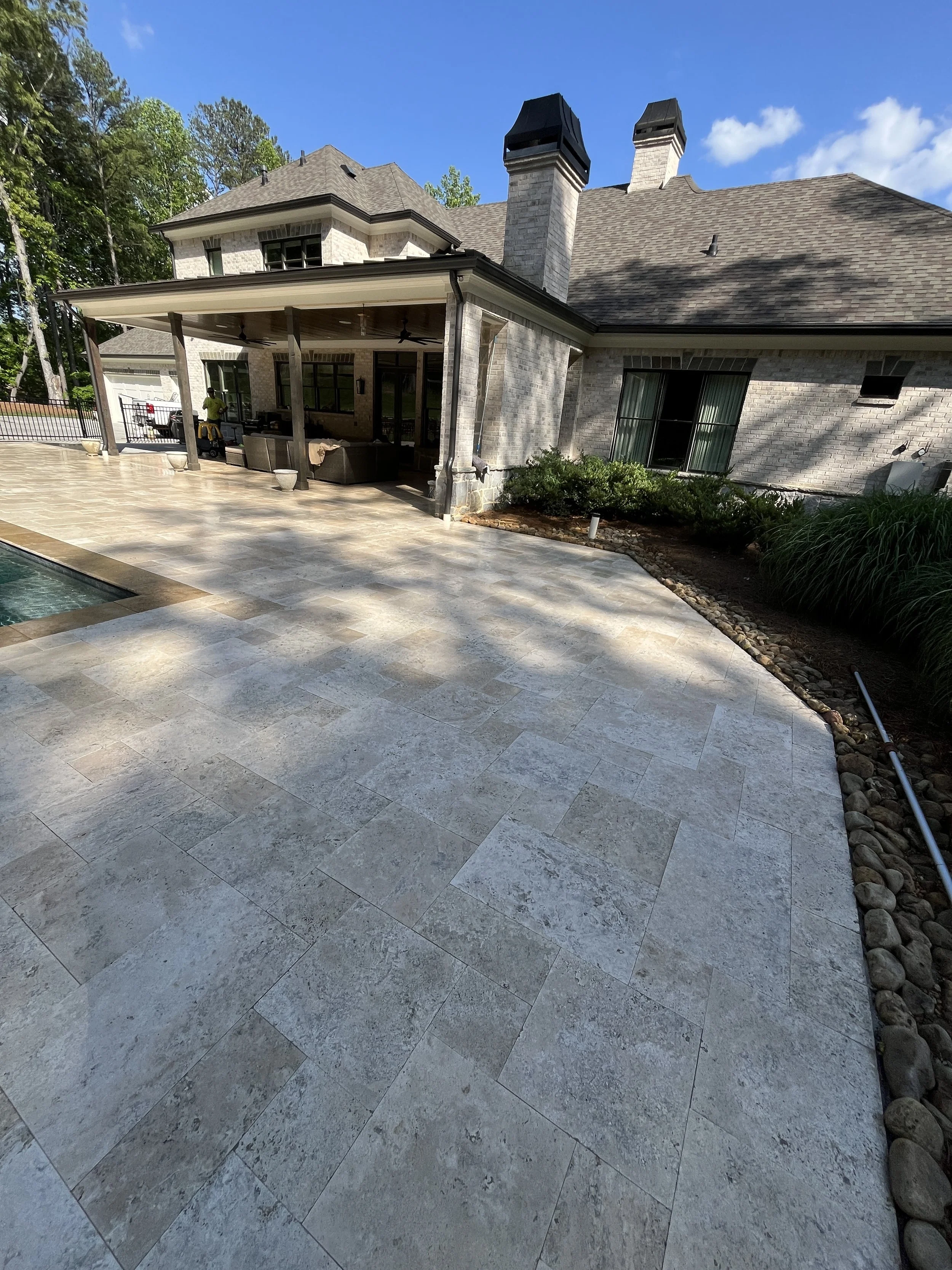 A backyard patio with large stone tiles, outdoor furniture under a covered porch, and a view of a house with multiple chimneys and windows, surrounded by trees and a blue sky.