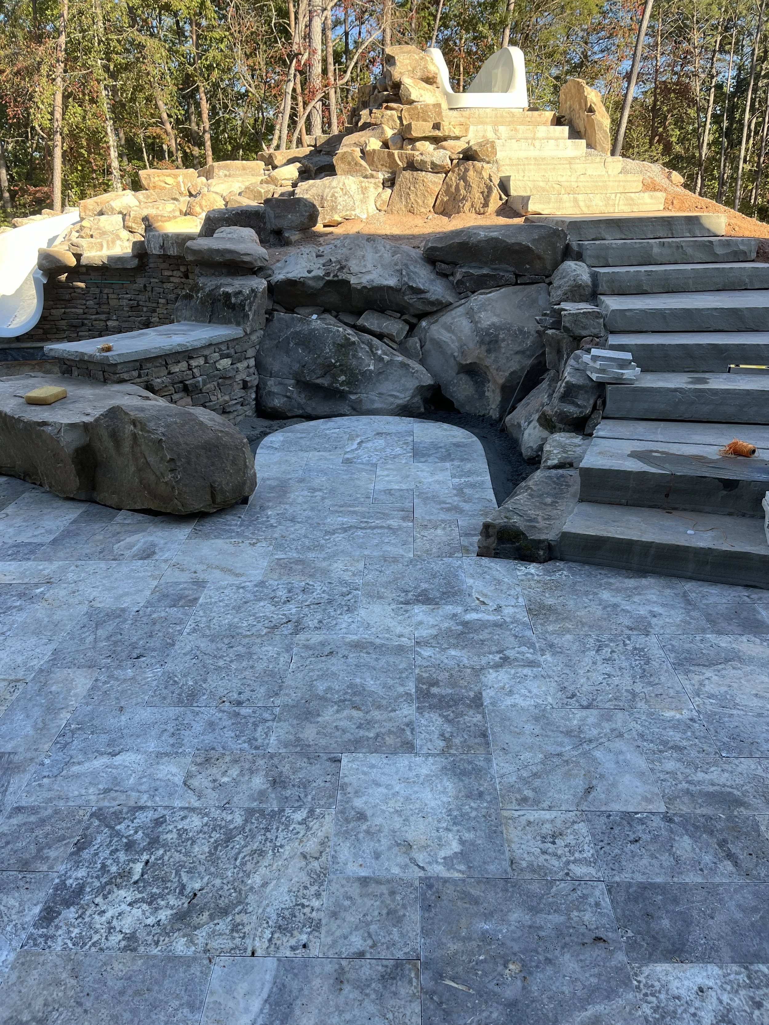 Outdoor scene of a stairway under construction, with stone steps and a slide in the background, surrounded by trees with fall foliage.
