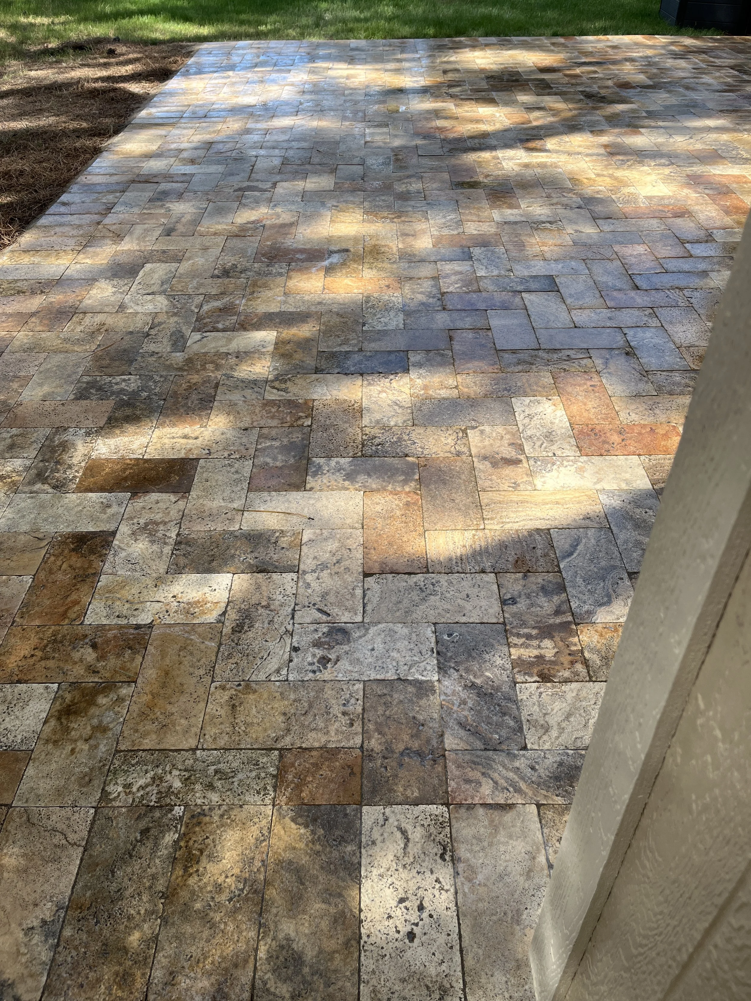 A rectangular stone patio with multicolored paving stones in shades of brown, beige, gray, and tan, partially shaded by nearby trees, with a grassy yard in the background.