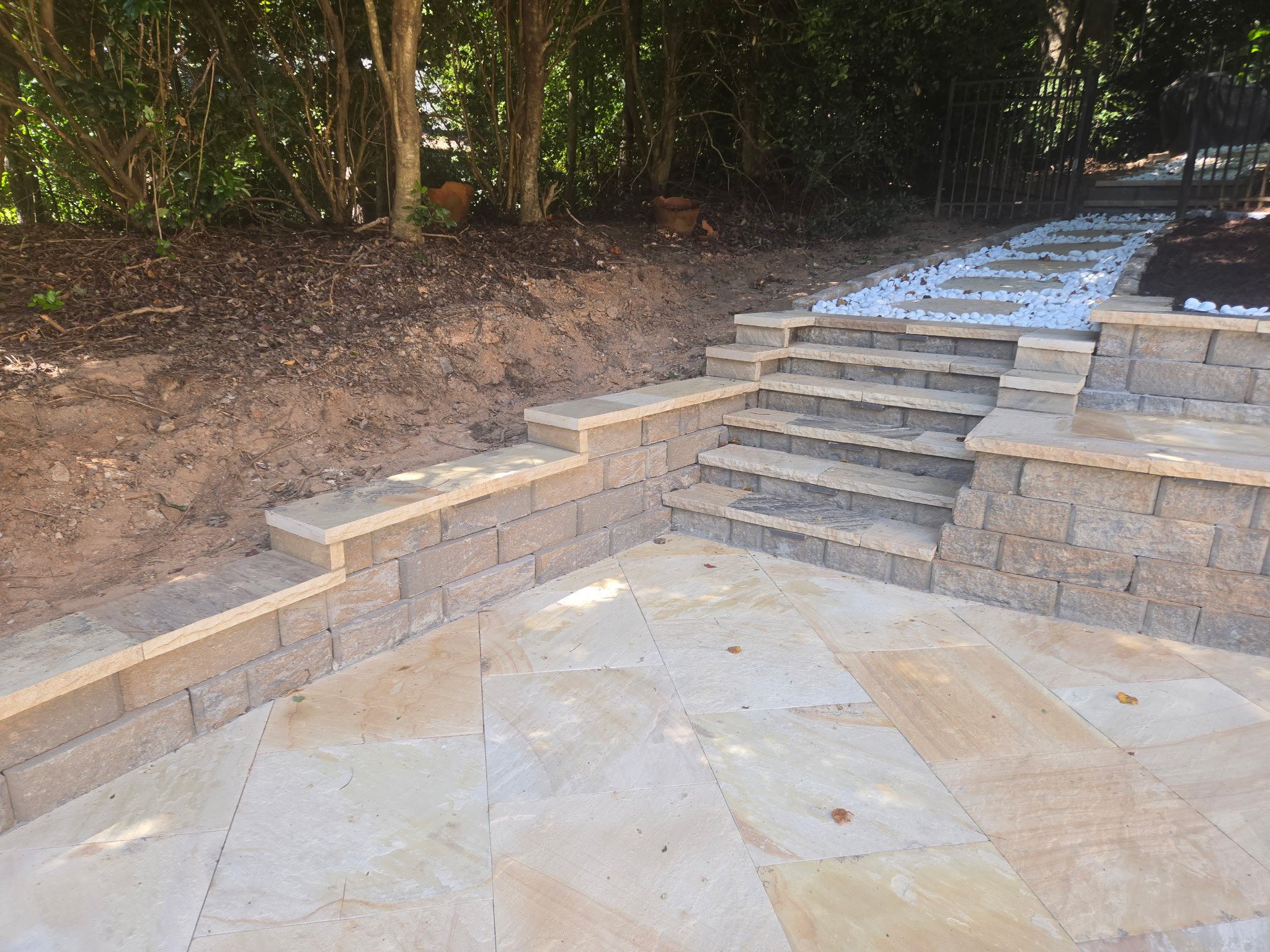 Newly constructed stone stairs with a curved design, leading up to a fenced area, bordered by a dirt slope with trees and greenery in the background.