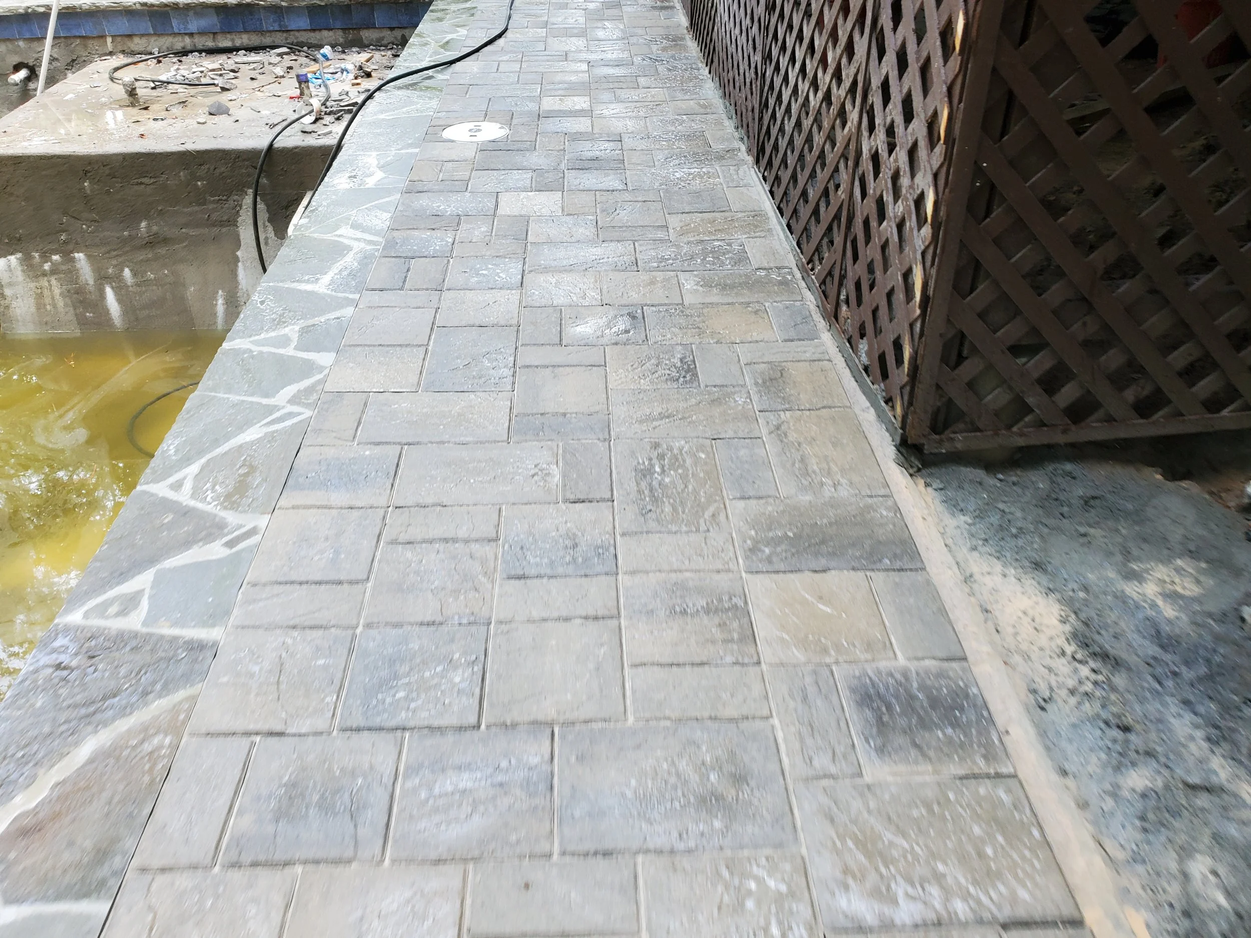 Concrete patio with square paver stones and a diamond pattern border, a fence on the right, and a construction area with tools and debris on the left.
