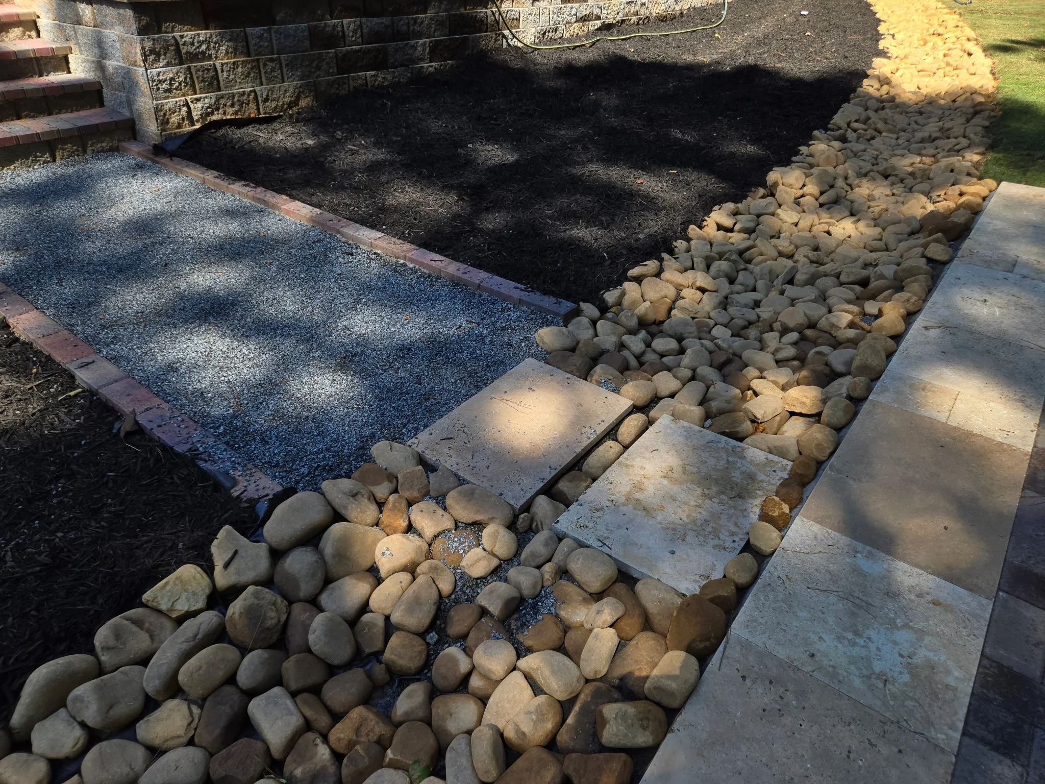 A garden walkway with a stone border, a small gravel pathway, and a section of dark soil with a border of large, rounded rocks next to a concrete sidewalk.