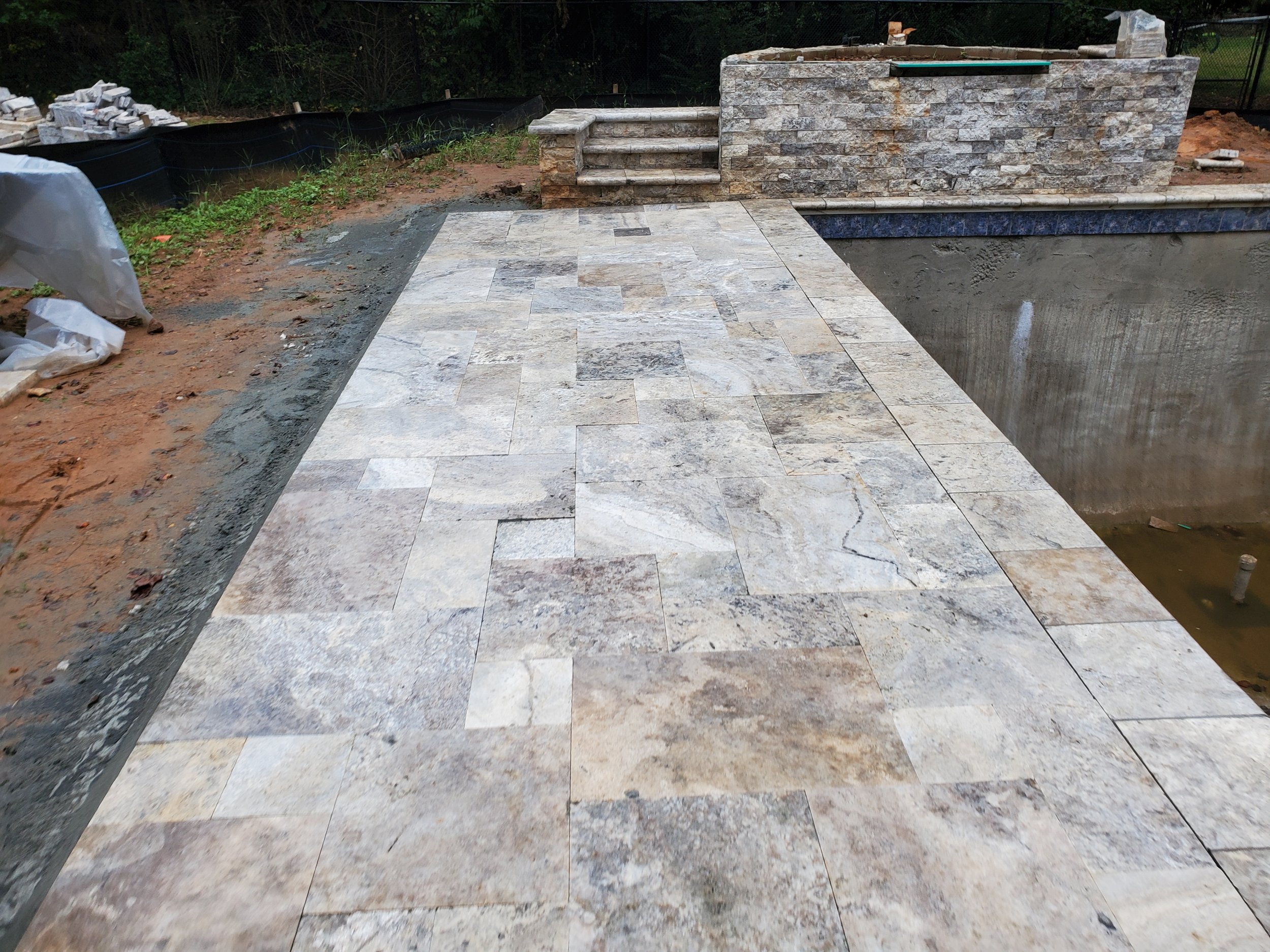 A partially constructed outdoor patio with stone tiles and a retaining wall under construction, surrounded by dirt and construction materials.