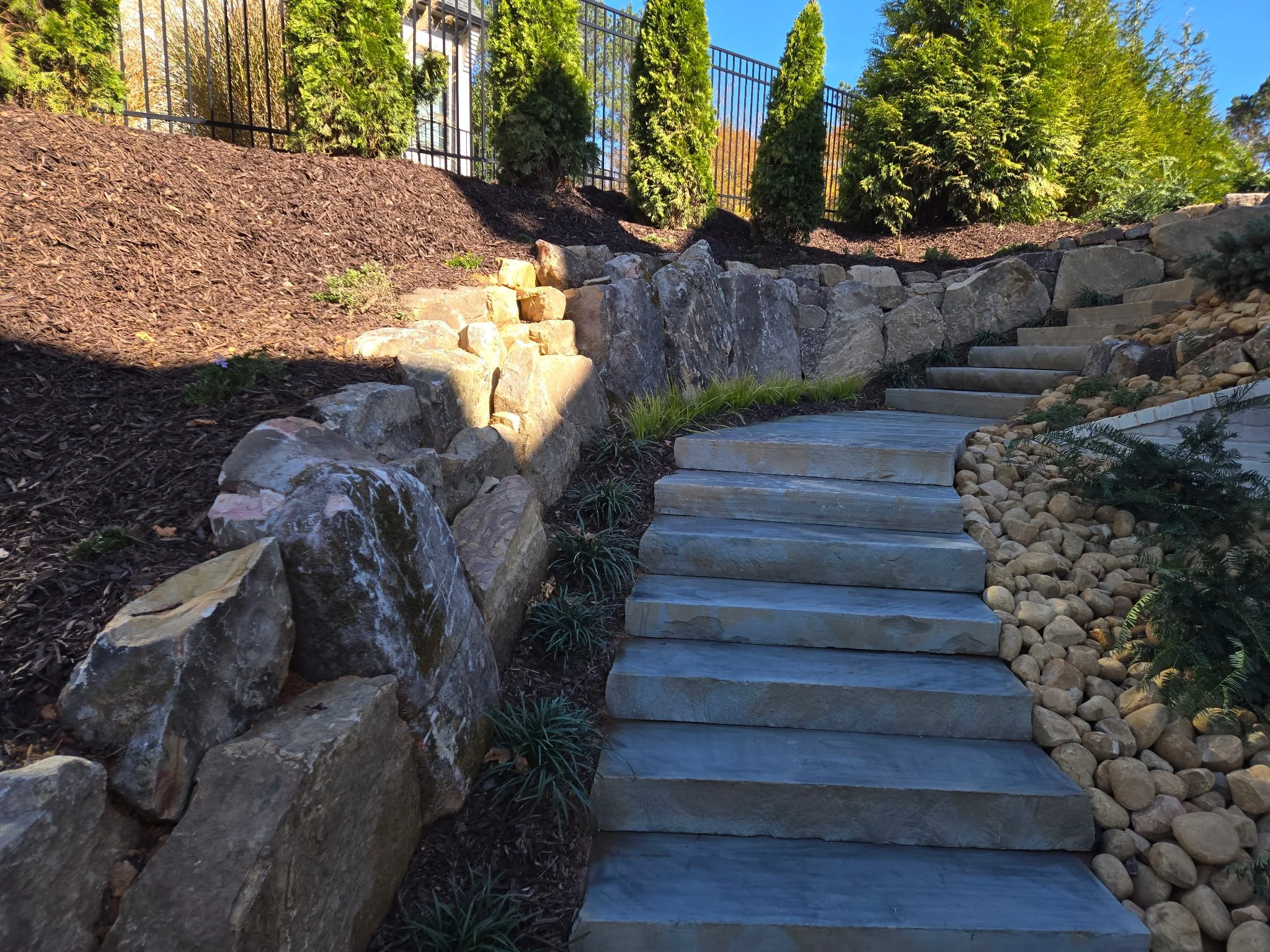 Stone steps leading uphill along a landscaped area with large rocks, small plants, mulch, and drought-tolerant bushes, with a black metal fence at the top and green trees in the background.