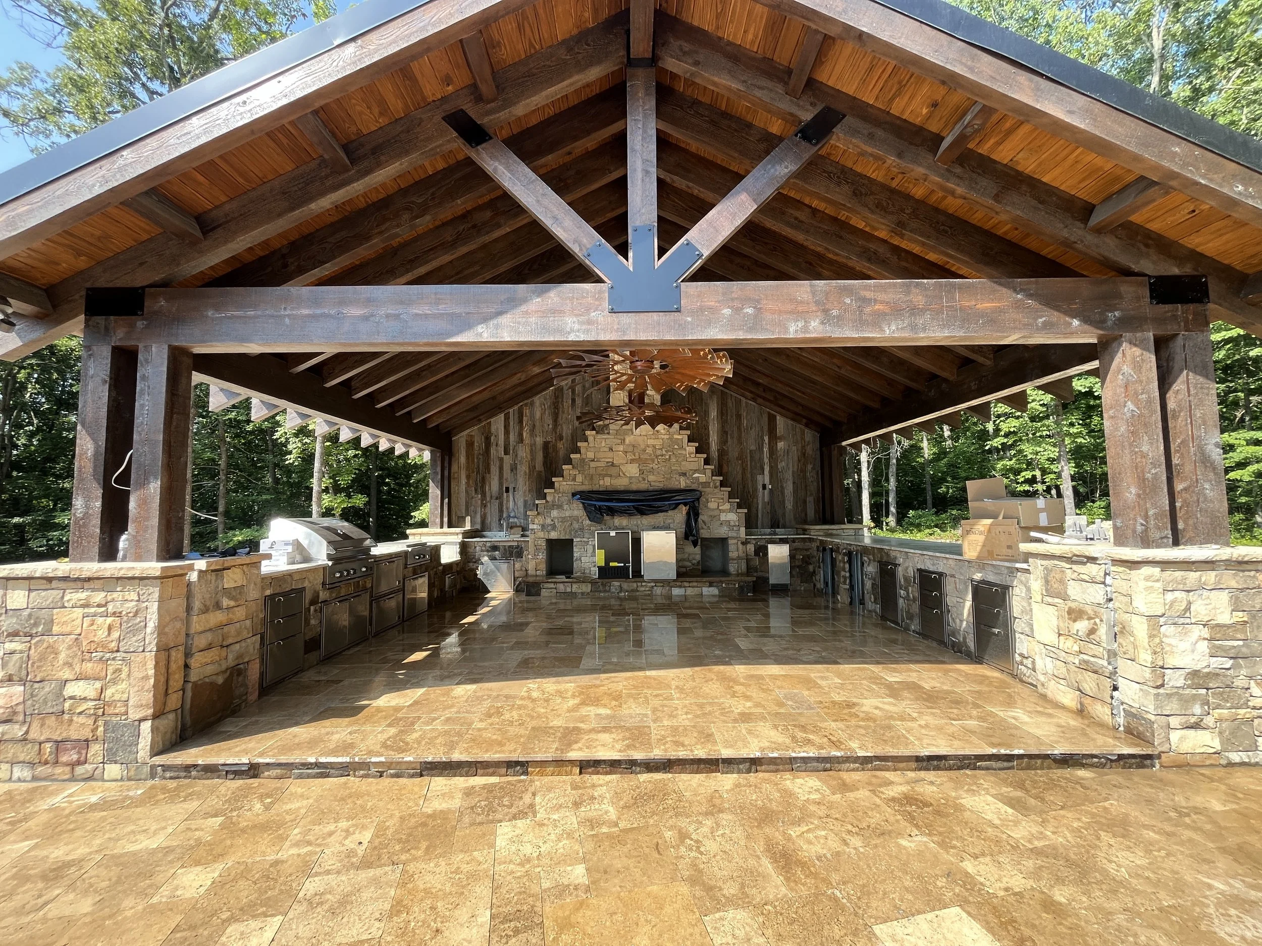 A spacious outdoor kitchen with a wooden roof supported by wooden beams, surrounded by trees. The kitchen has a stone countertop with multiple stainless steel appliances, a built-in grill, and a brick fireplace in the background. The floor is tiled w