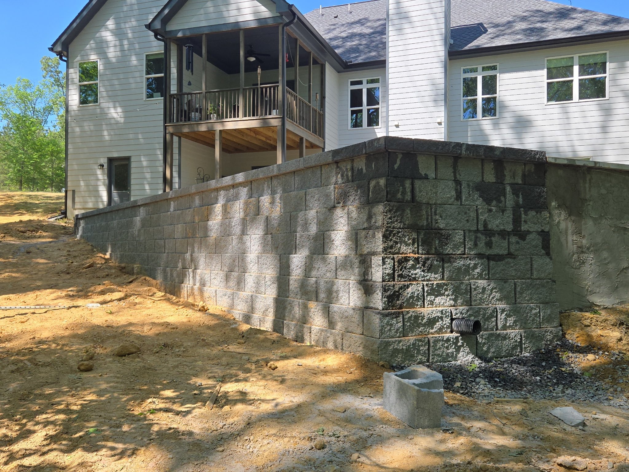 Backyard of a house with a partially constructed balcony and a stone retaining wall, surrounded by trees and a clear blue sky.