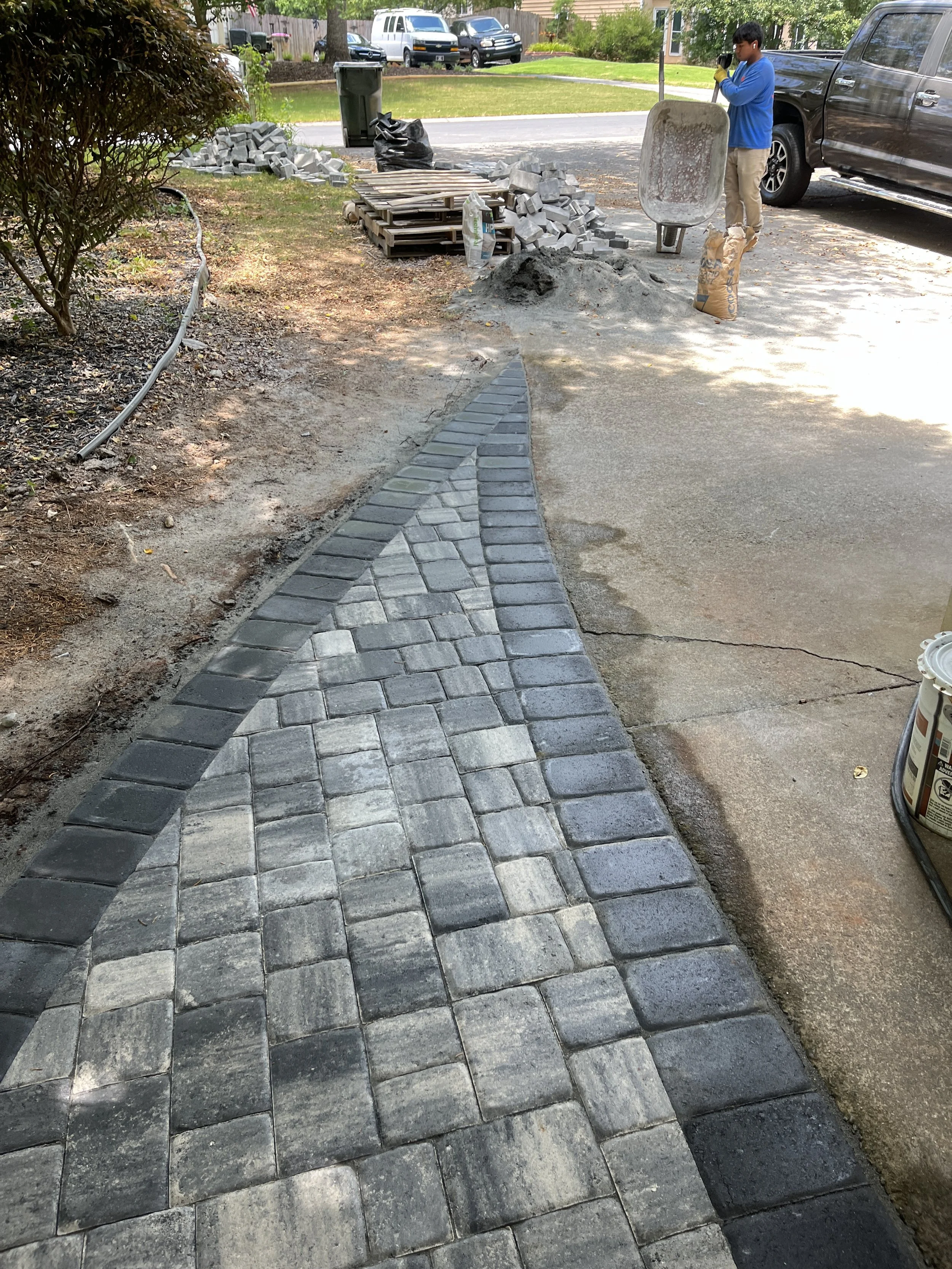 A person installing a brick sidewalk or pathway in a residential yard next to a mud and dirt area. There are stacks of bricks, a wheelbarrow, and some construction tools nearby, with a black pickup truck parked on the street.
