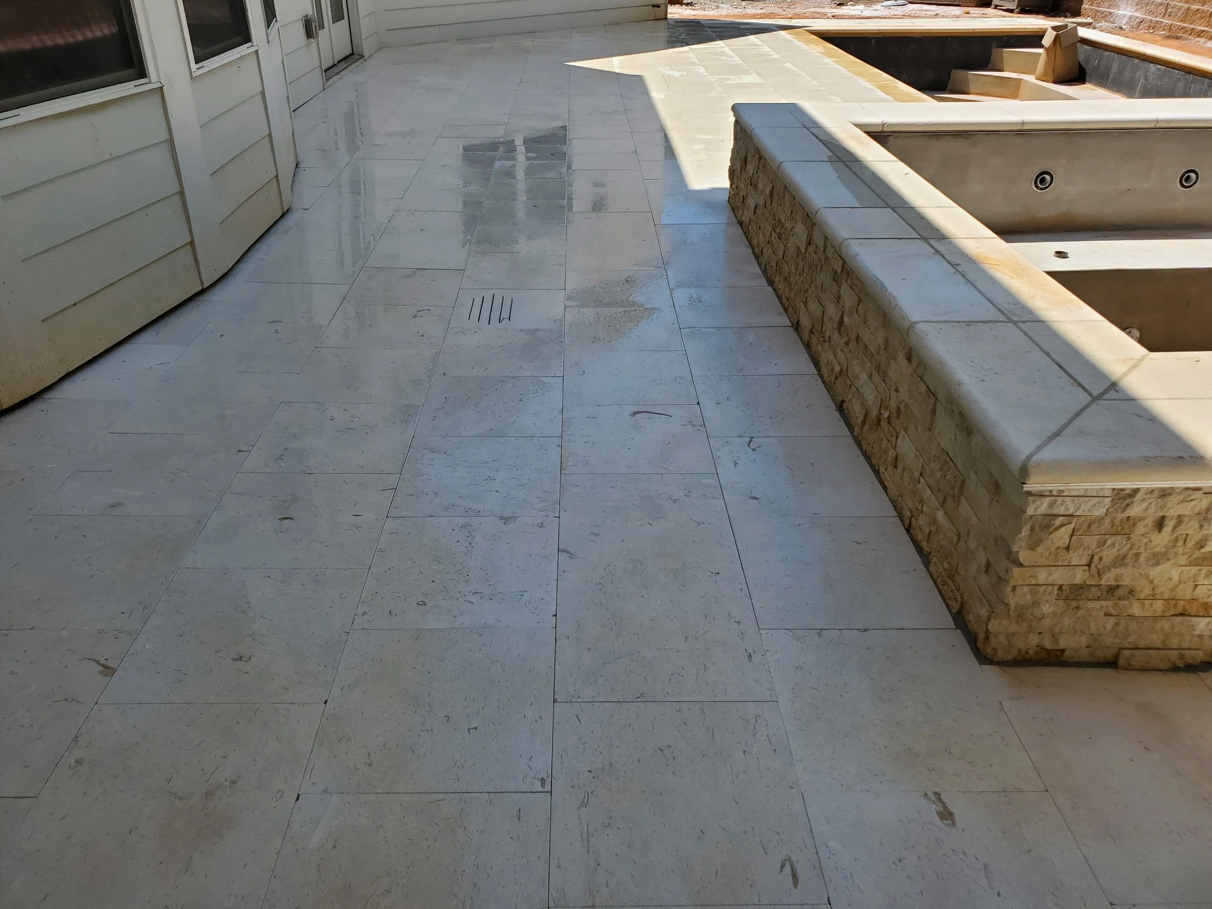 An outdoor patio with beige stone tiles on the ground and a built-in corner bench with stone and brickwork. Part of the patio is shaded, and there are windows and a door on the left side.