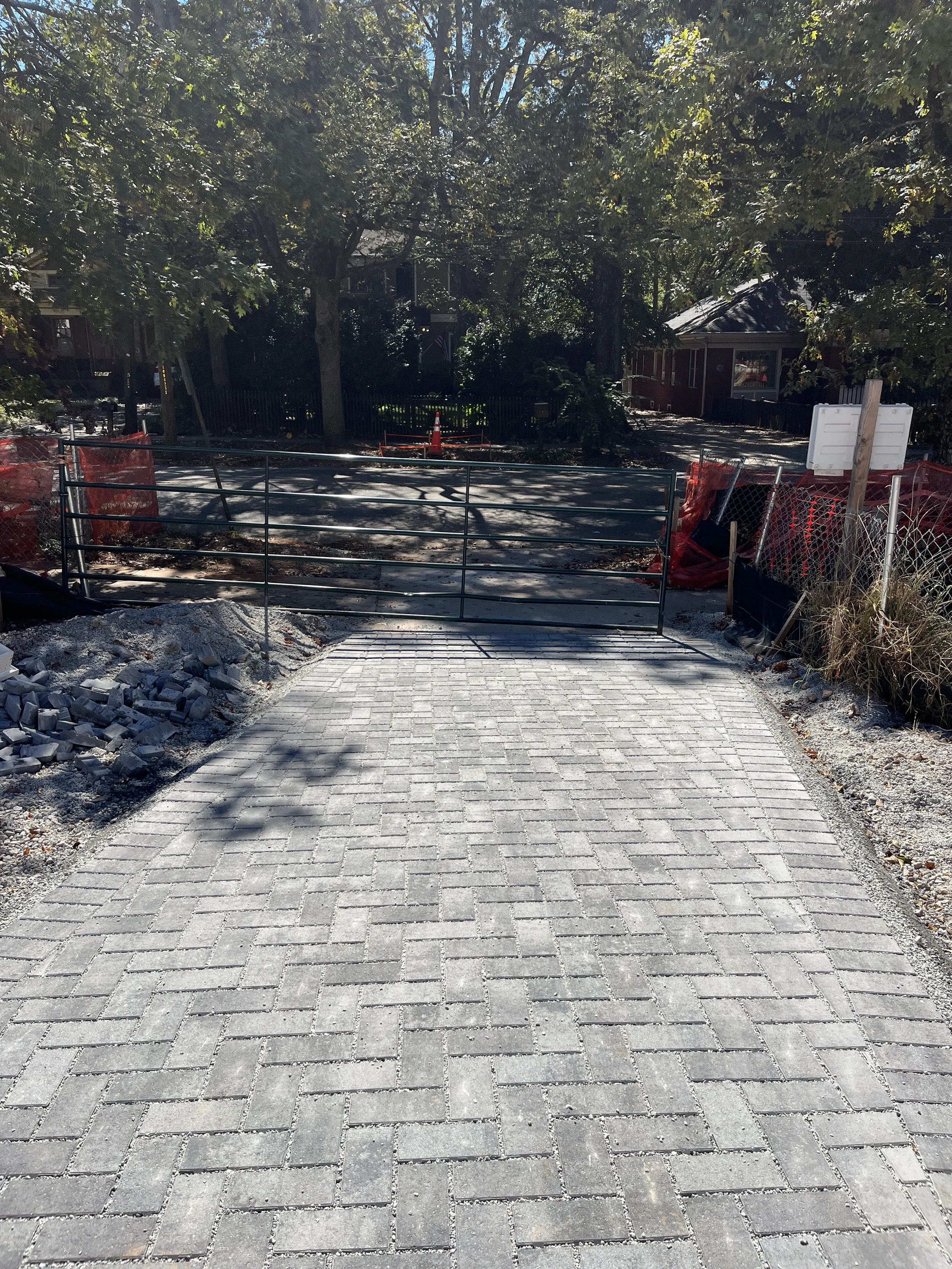 A sidewalk under construction with paving stones arranged in a herringbone pattern, surrounded by construction safety barriers, fencing, and trees with green leaves, with a house in the background.