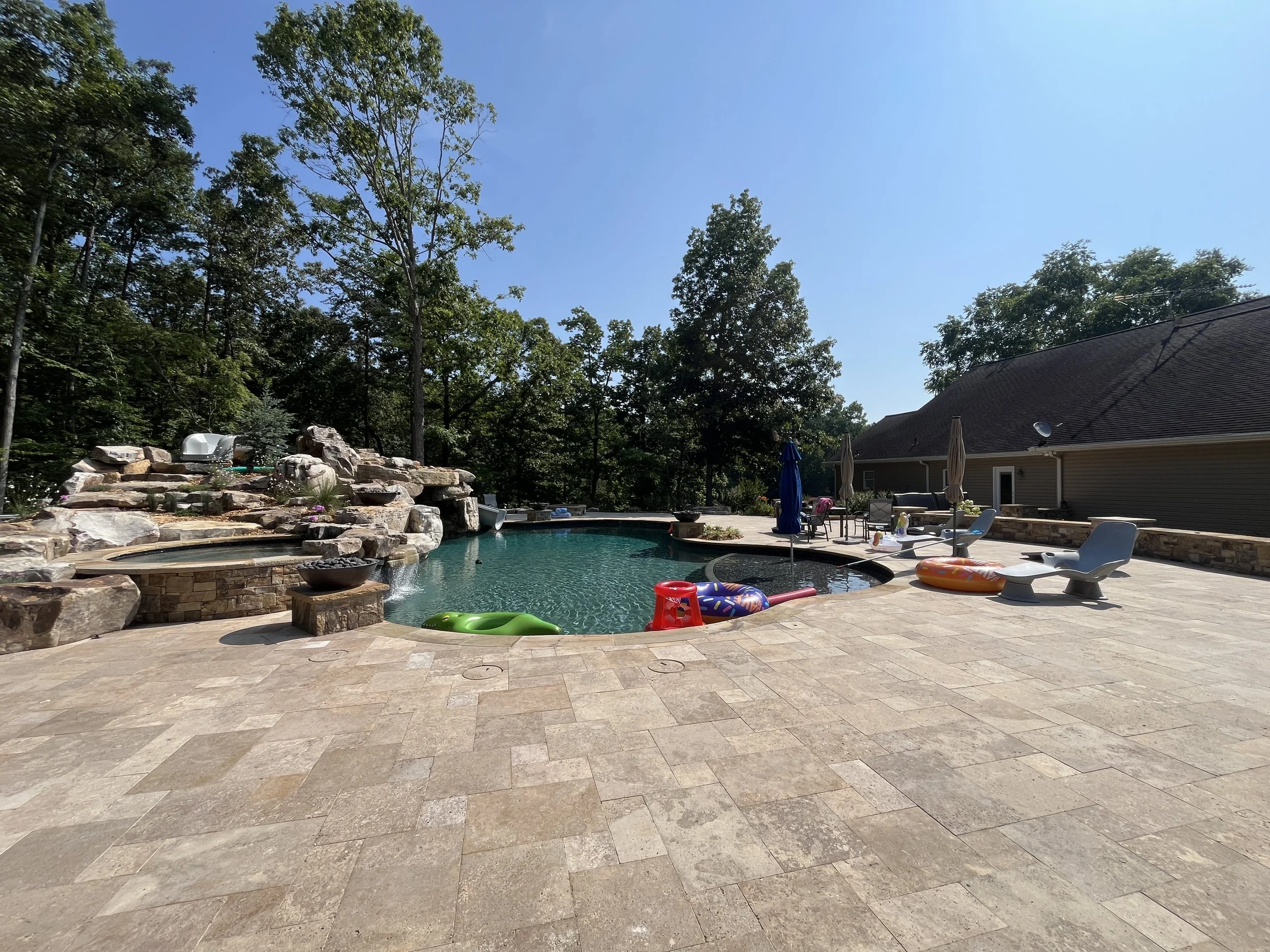 Backyard with swimming pool, patio furniture, umbrellas, and trees under a clear blue sky.