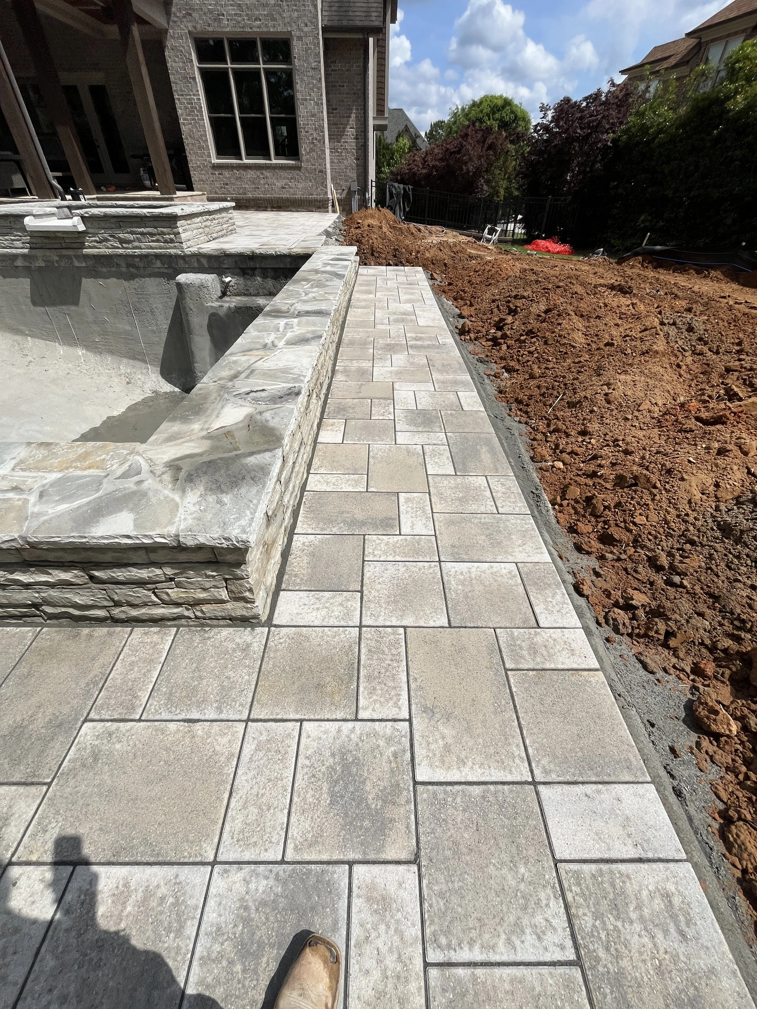 Construction site with partially completed stone patio and walkway, dirt and construction materials on the side, and a house in the background.