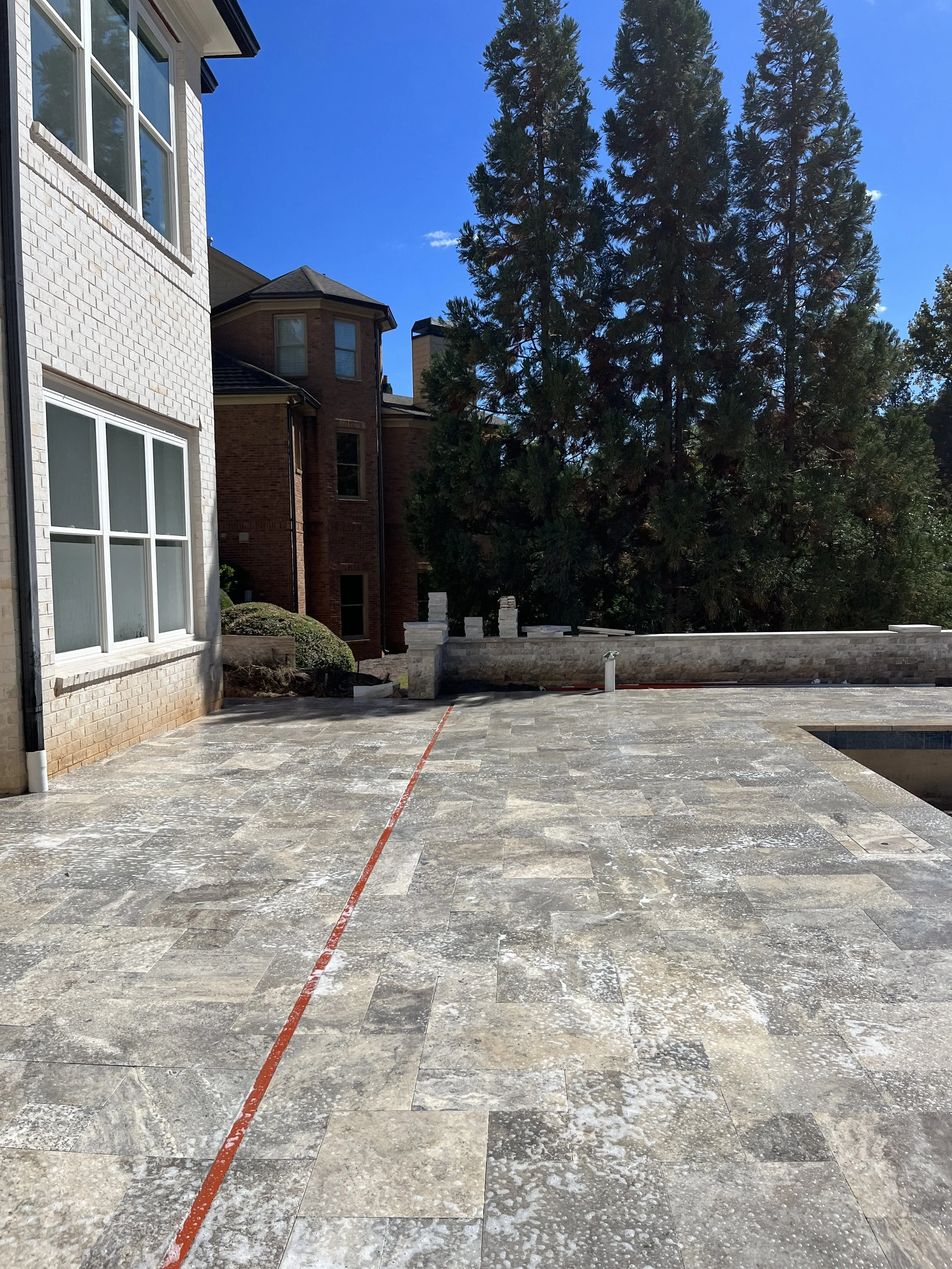 View of a backyard patio with tiled flooring, a modern house with large windows on the left, a tall pine tree in the background, and neighboring houses with brick facades, under a clear blue sky.