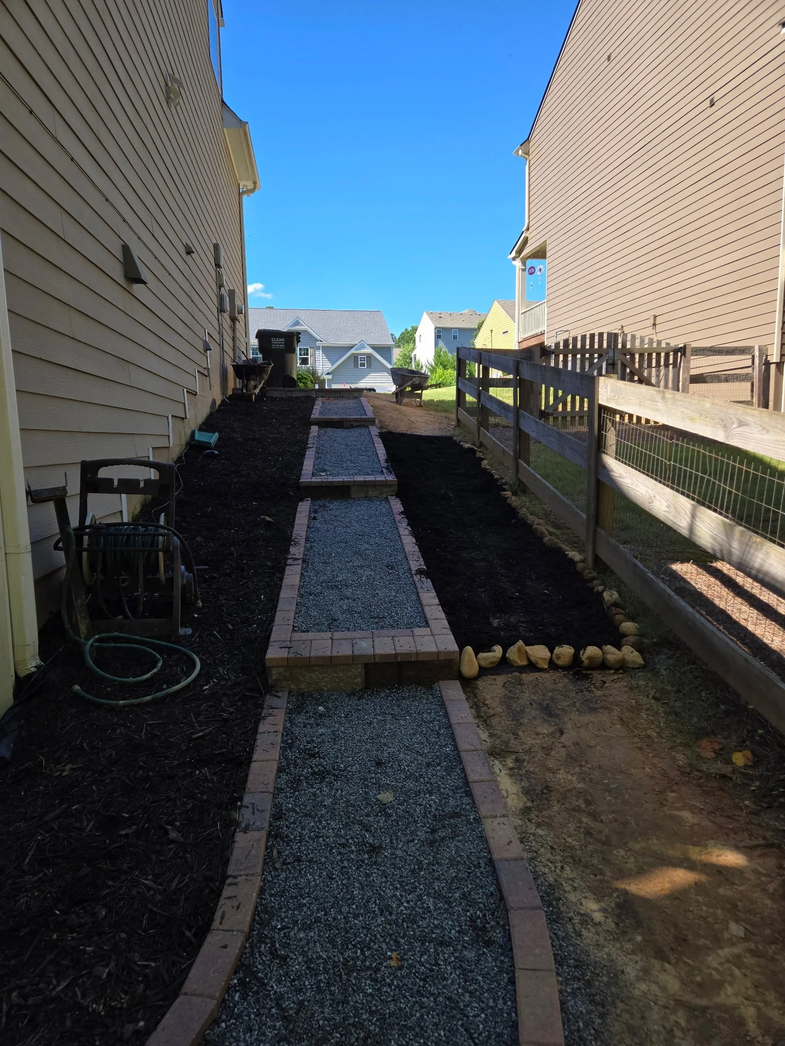 A backyard pathway under construction with a gravel walkway bordered by bricks, on a sunny day with clear blue sky and neighboring houses visible in the background.