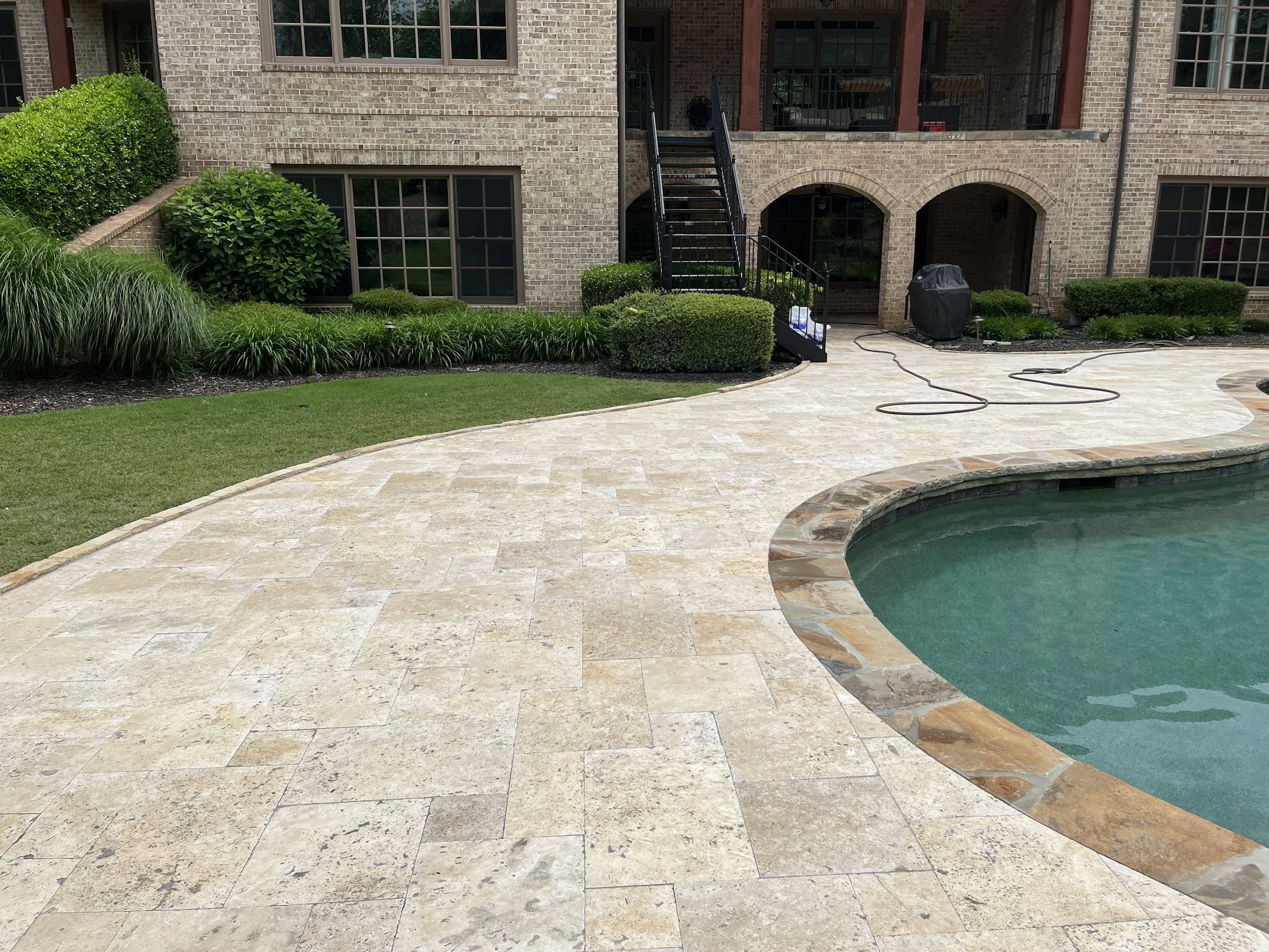 Backyard patio with stone paving, swimming pool, lush greenery, bushes, and a brick house with balconies and stairs.