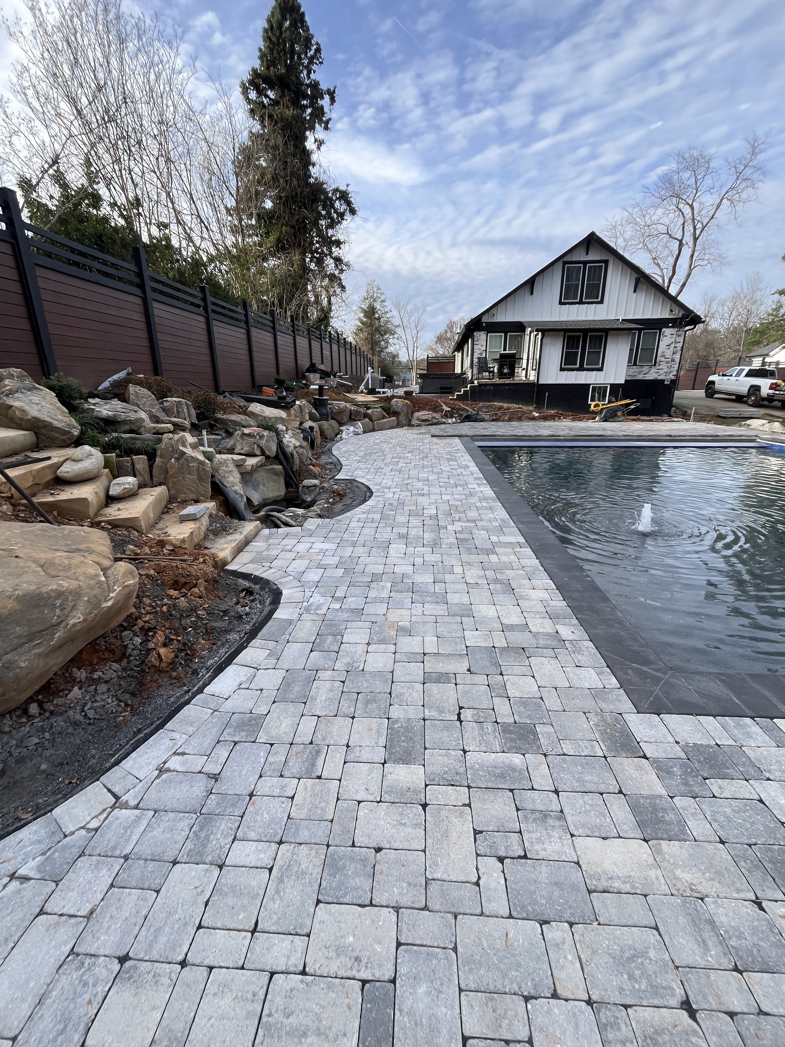 A backyard with a newly paved stone patio and a pool, surrounded by a low rock wall and a tall fence, with a house in the background. The yard is under landscaping with some tools and construction materials visible.