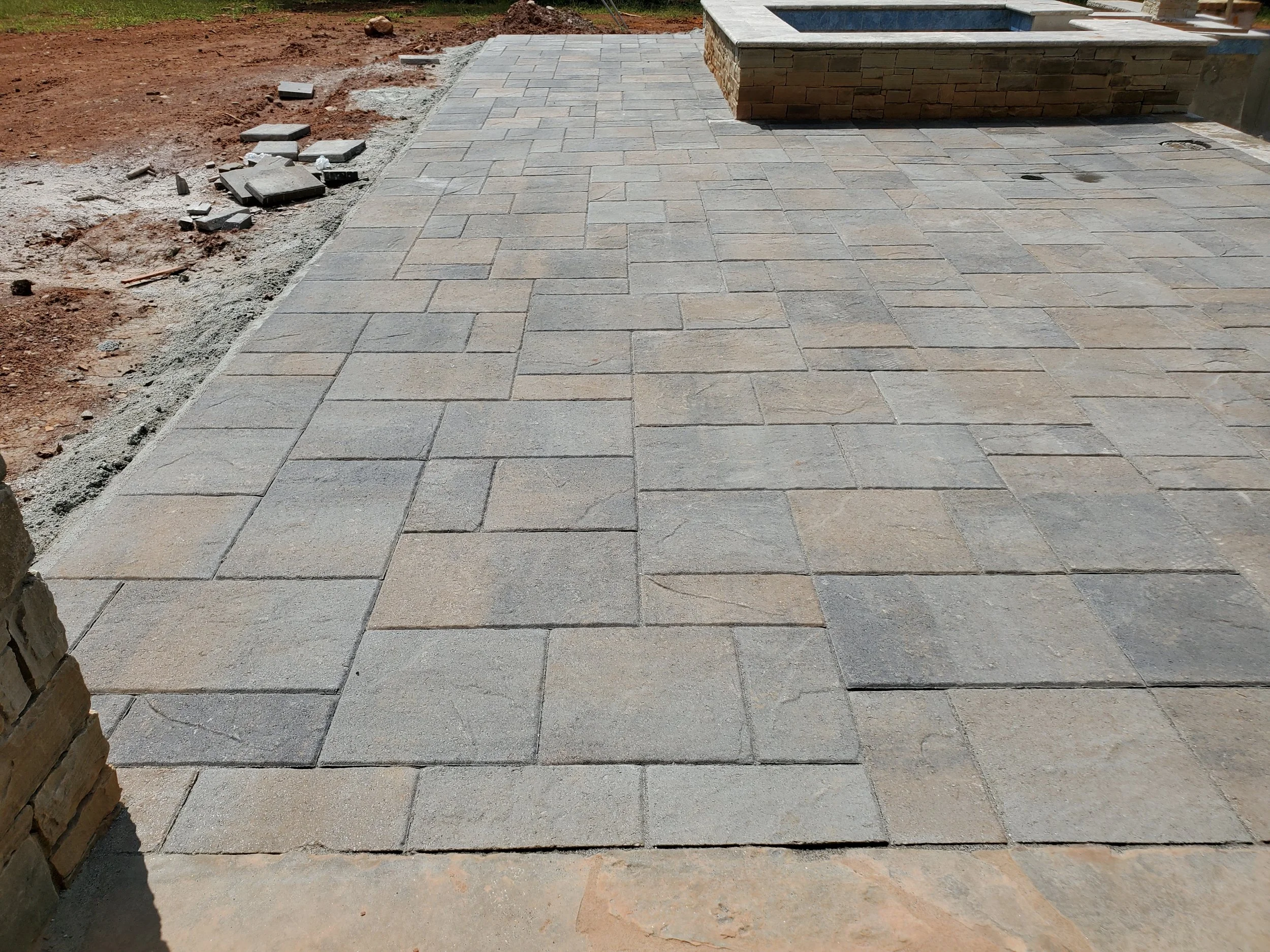 The image shows a newly paved patio area with interlocking concrete pavers in shades of tan and gray. There is a raised fire pit or planter made of stone on the right side of the patio. On the left side, a section of dirt and uneven ground is visible