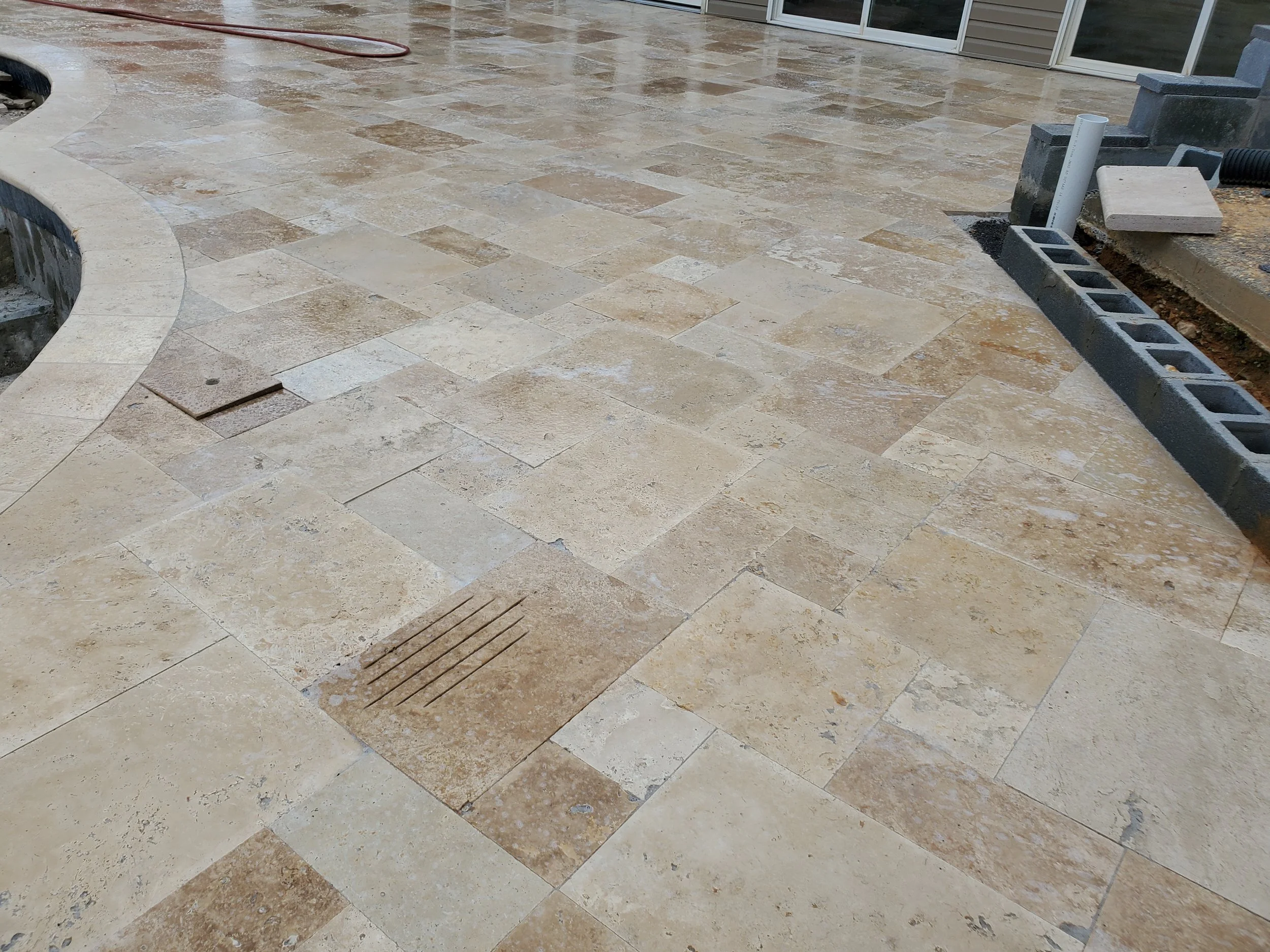 A newly constructed outdoor patio with beige and tan stone tiles, partially finished with some construction materials and tools nearby.