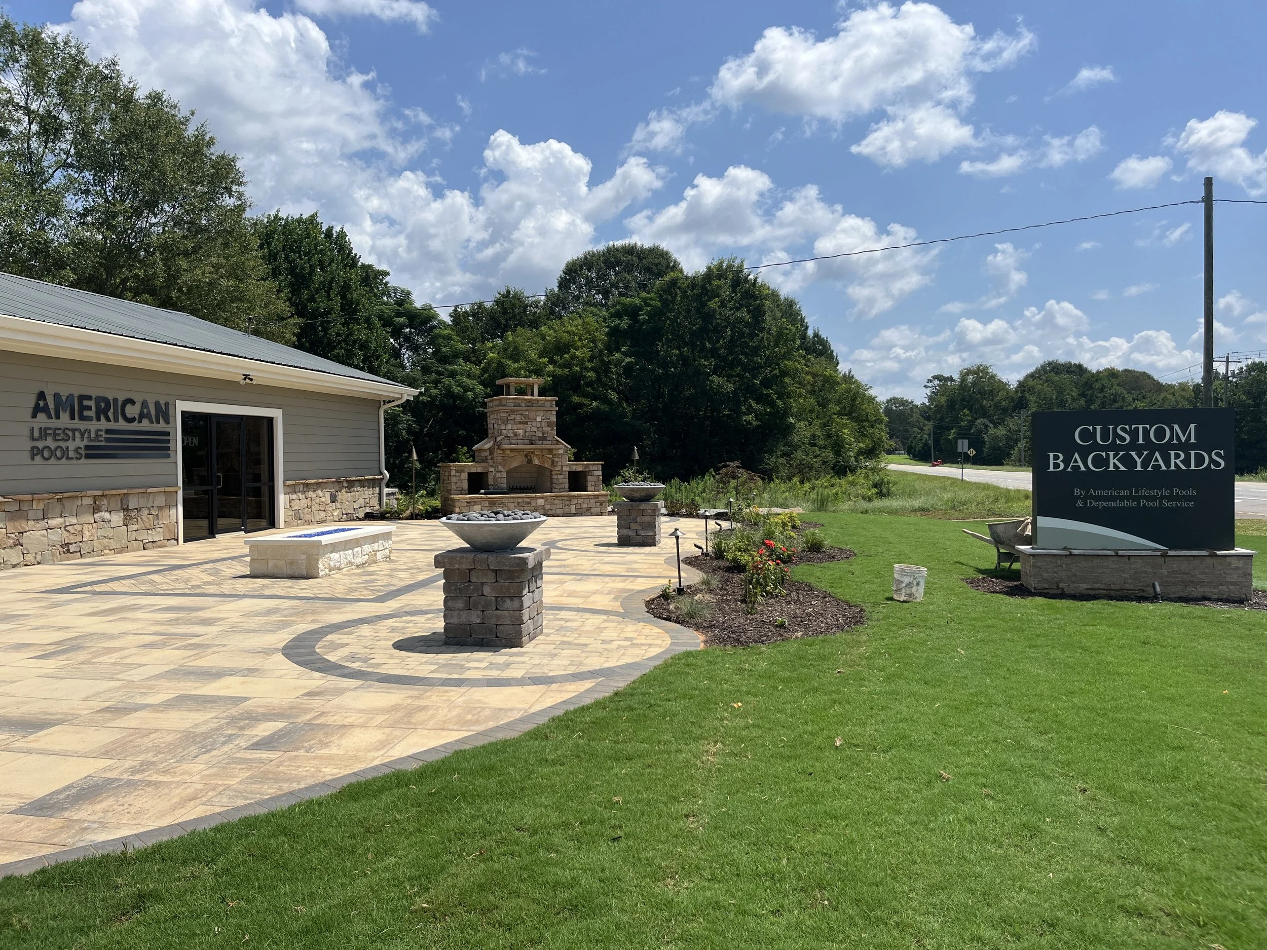 An outdoor patio area with a stone firepit, decorative planters, and a building with a sign that reads 'American Lifestyle Pools'. There is a grassy lawn, trees, and a blue sky with scattered clouds.