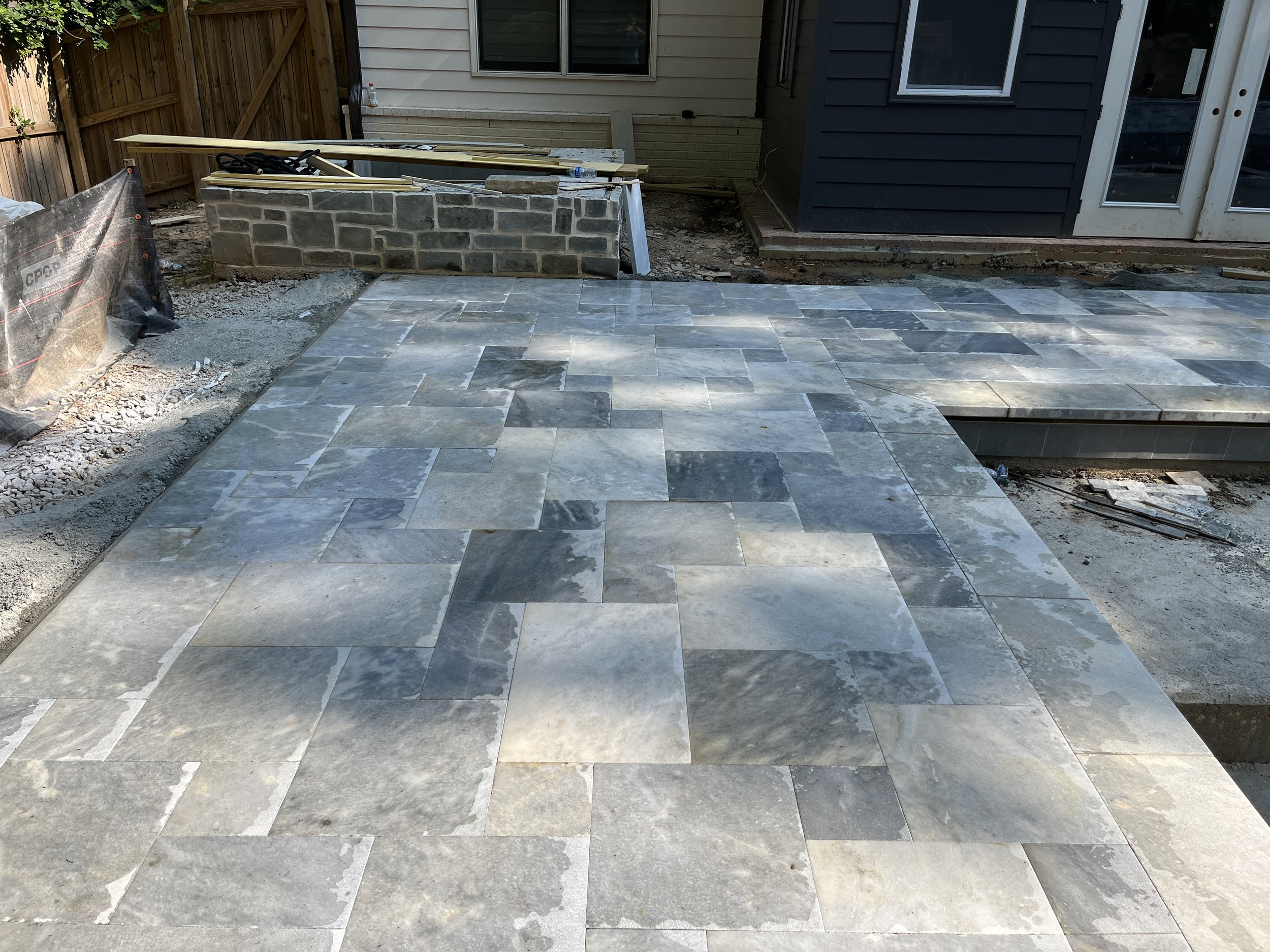 Backyard patio under construction with stone tiles, wooden fence, house exterior, and construction materials.
