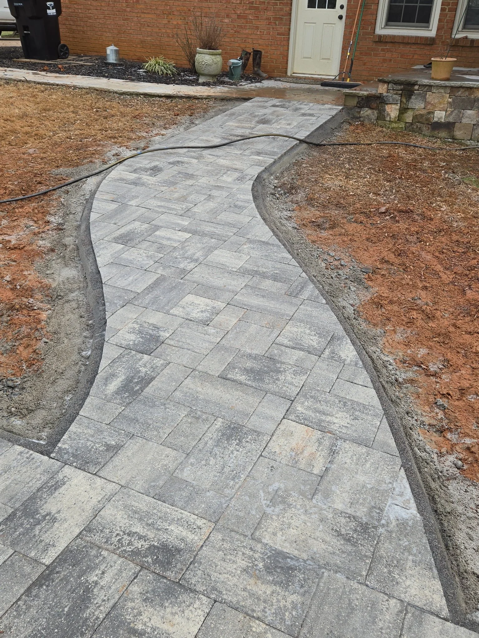 A new stone walkway is being installed in the yard of a house, with some areas already completed and others still under construction. The yard has dirt and patches of grass around the walkway.