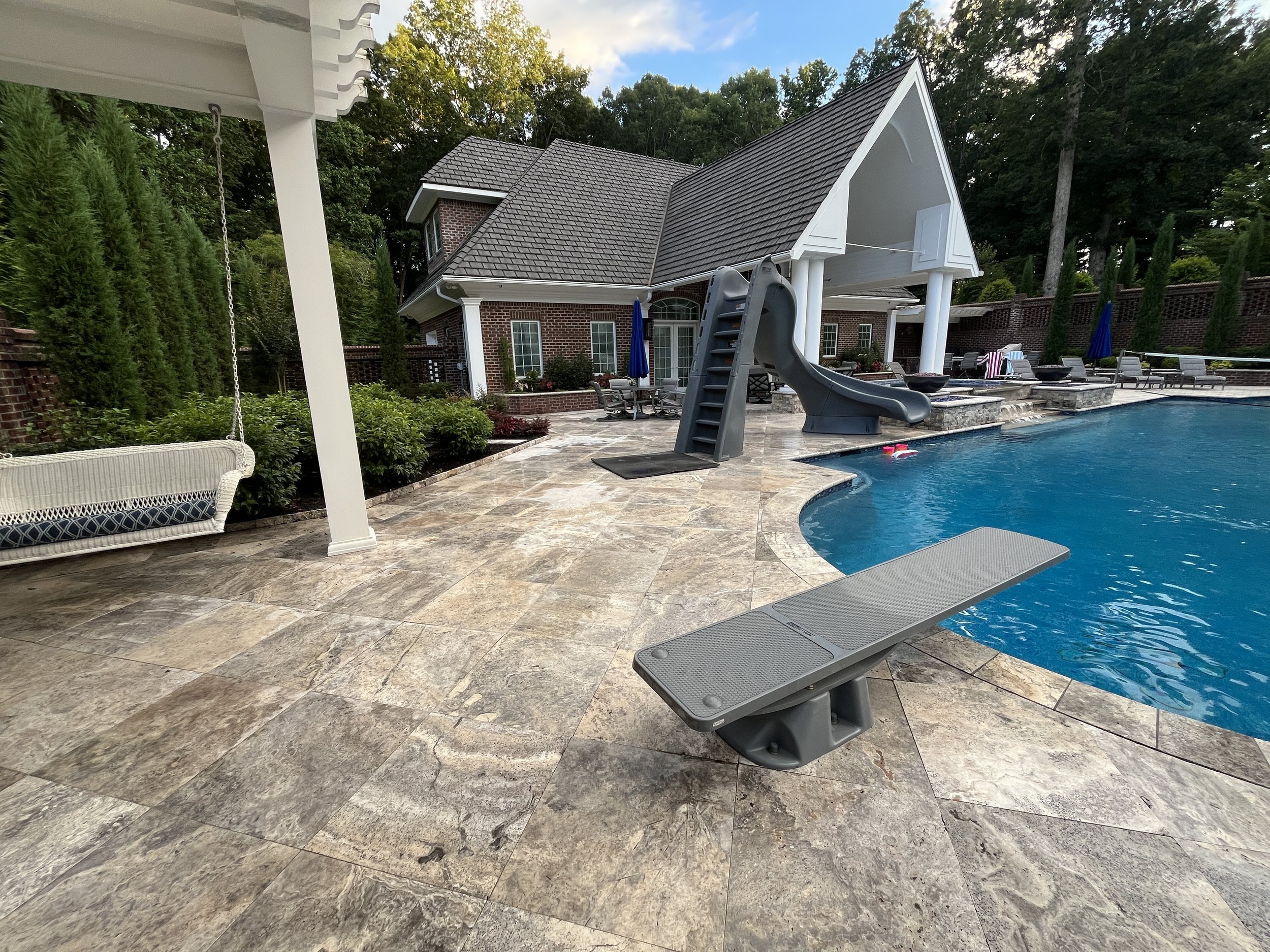 A backyard with a swimming pool, poolside lounge chairs, a small slide, a patio area with tables and chairs, a house with a brick exterior, and tall trees in the background under a partly cloudy sky.