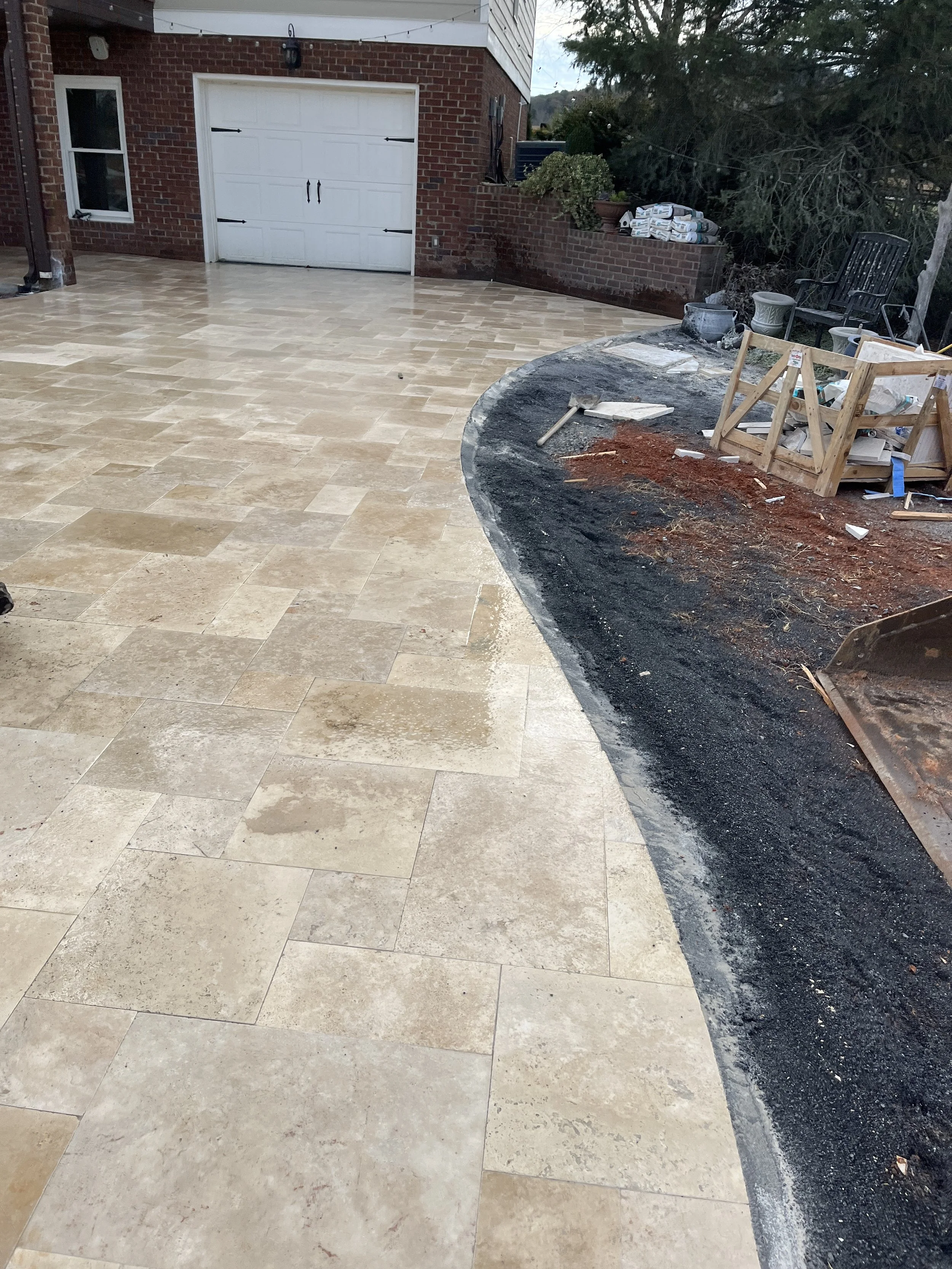 A backyard patio area with beige stone tiles, partially covered by construction materials and tools, and a garden bed with some plants and stacked bags of materials, with a garage door and brick wall in the background.