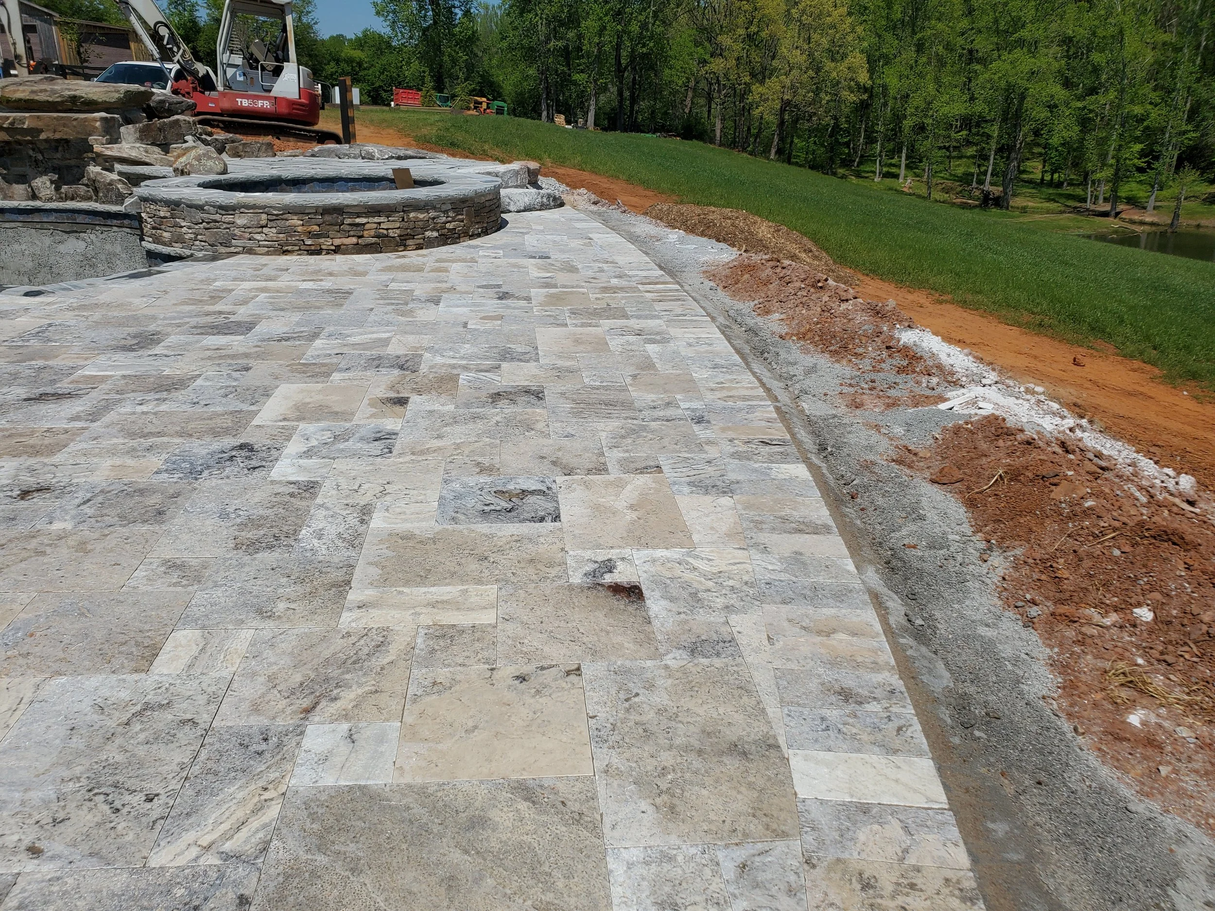 A construction site with freshly laid stone tiles forming a patio, a stone firepit, and a small explosive-looking device on the firepit. In the background, there are trees and construction equipment, including a small excavator and piles of earth.
