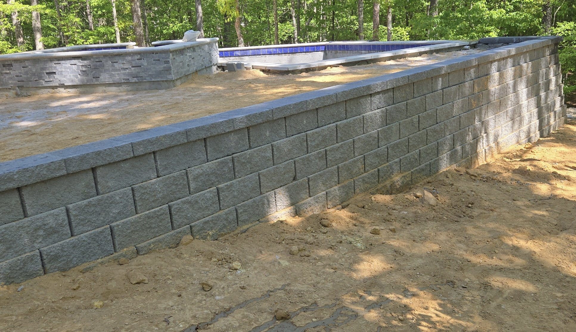 Partially constructed outdoor pool with brick and stone walls, set in a wooded area, with visible dirt and construction materials around.