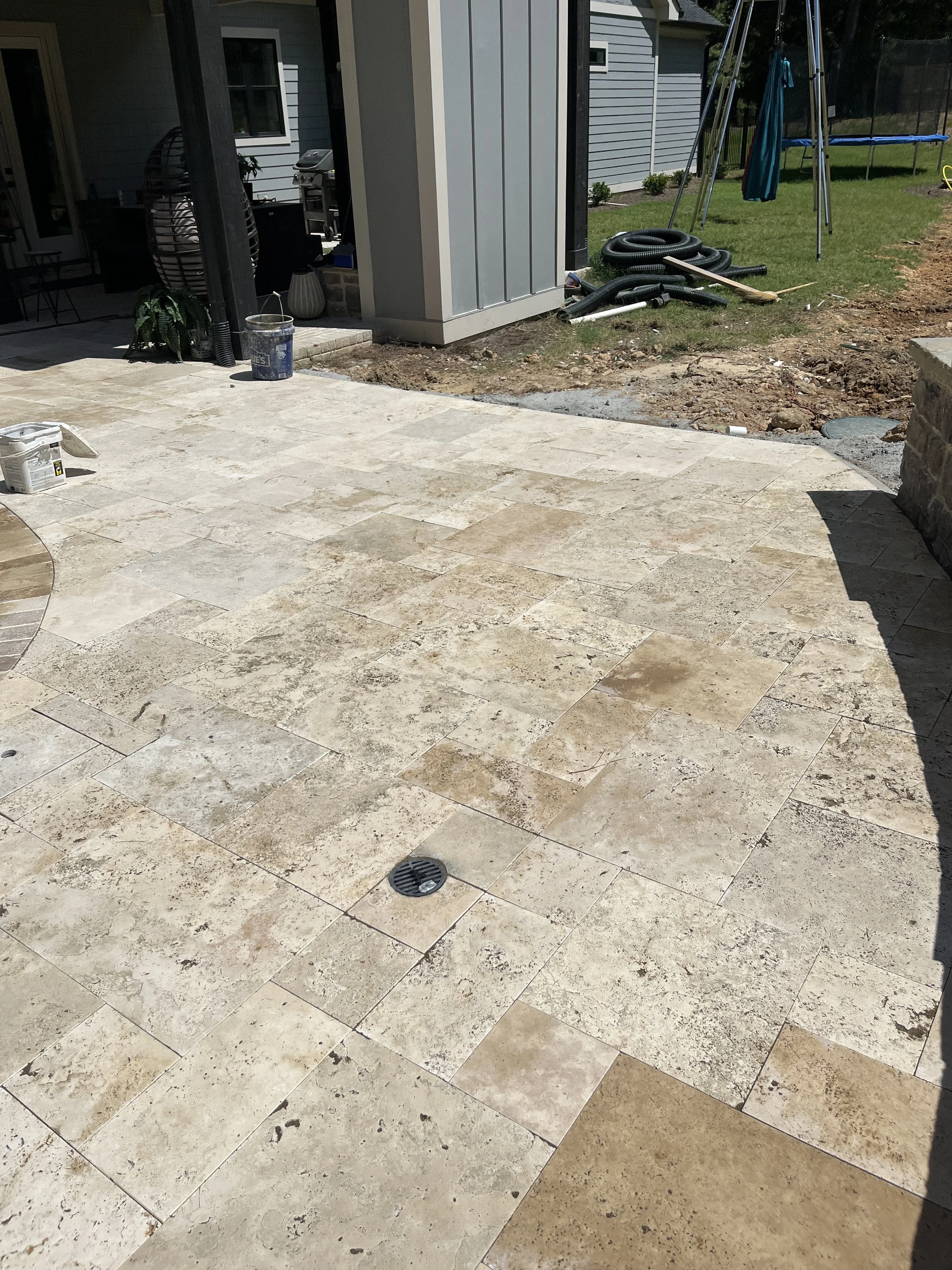 Backyard patio with beige tile flooring, construction materials and tools, including a bucket, pipes, and a pole, with a grassy yard, trampoline, and a house in the background.