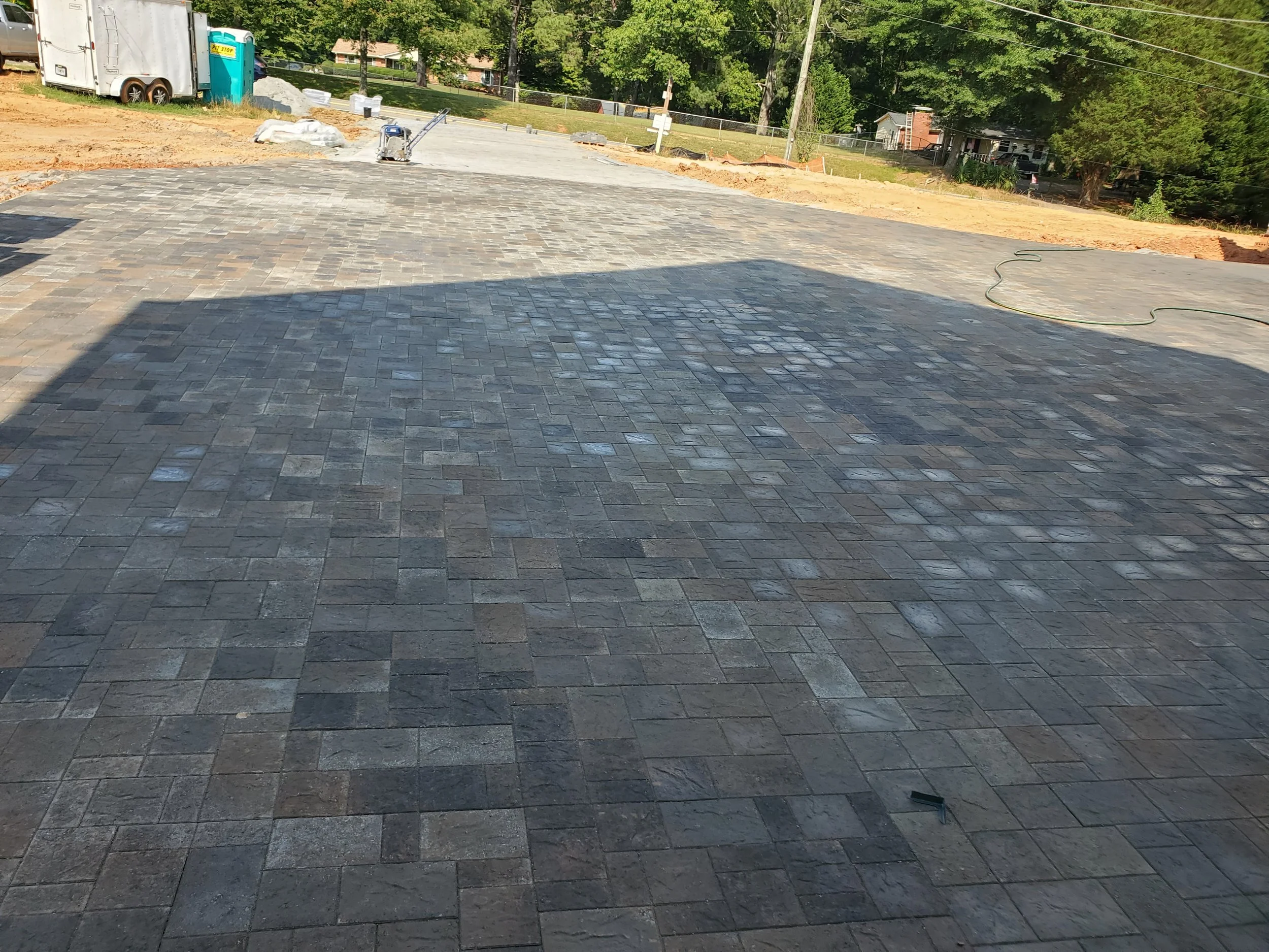 Newly paved stone patio or driveway with outdoor construction tools and materials nearby, in a residential backyard with trees and houses in the background.