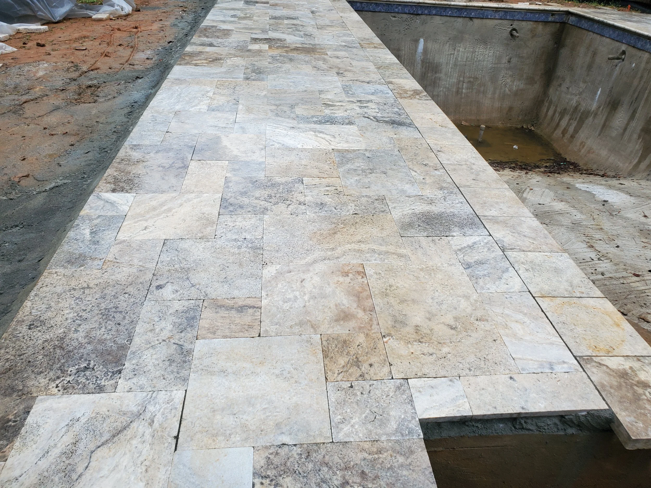 Photo of a stone tile walkway in a pool area under construction, with an empty swimming pool and construction materials around.