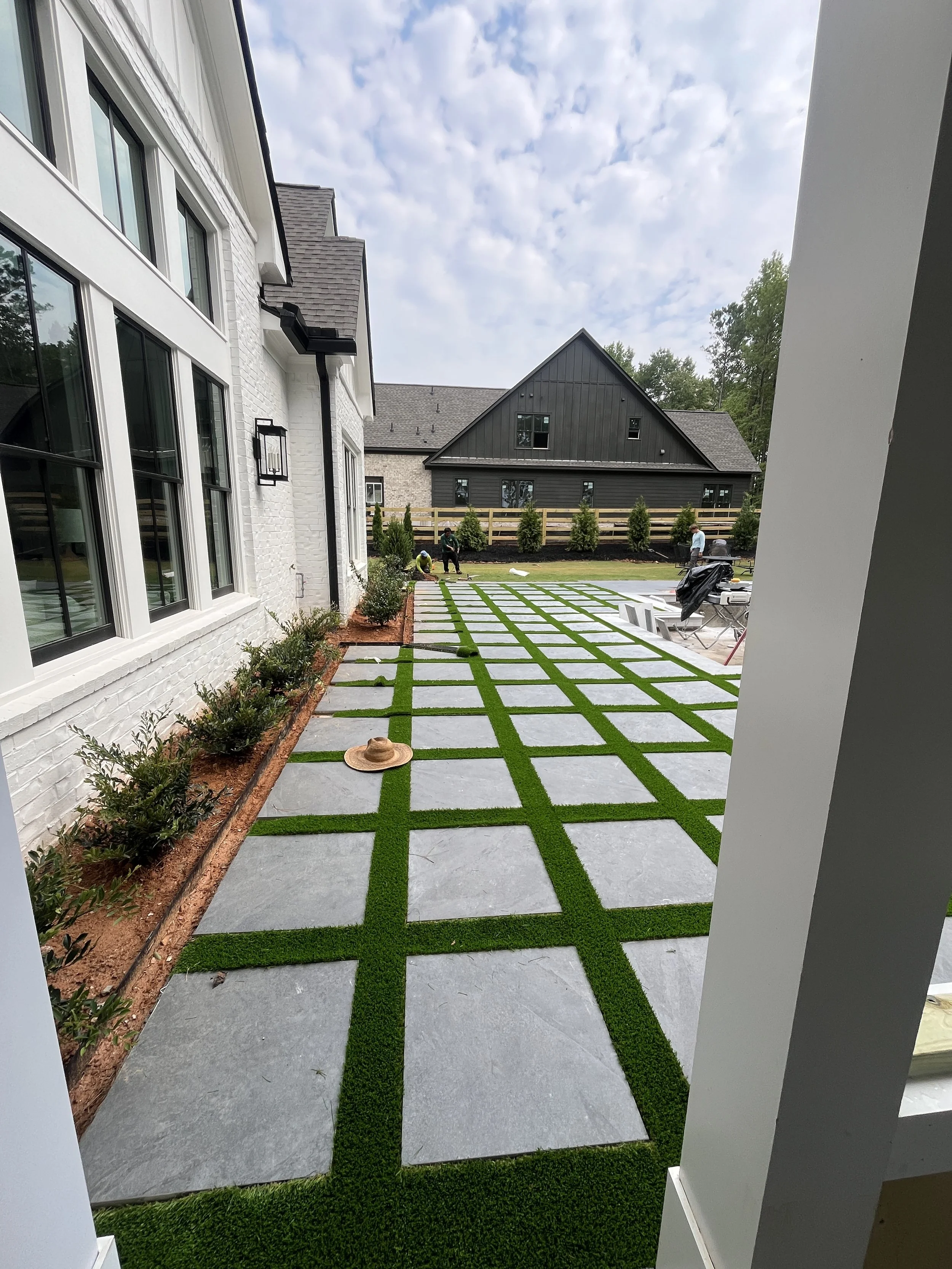 View of a backyard with a patterned concrete and artificial grass lawn, a flower bed along a white brick house, several workers installing the lawn, and a black house in the background.