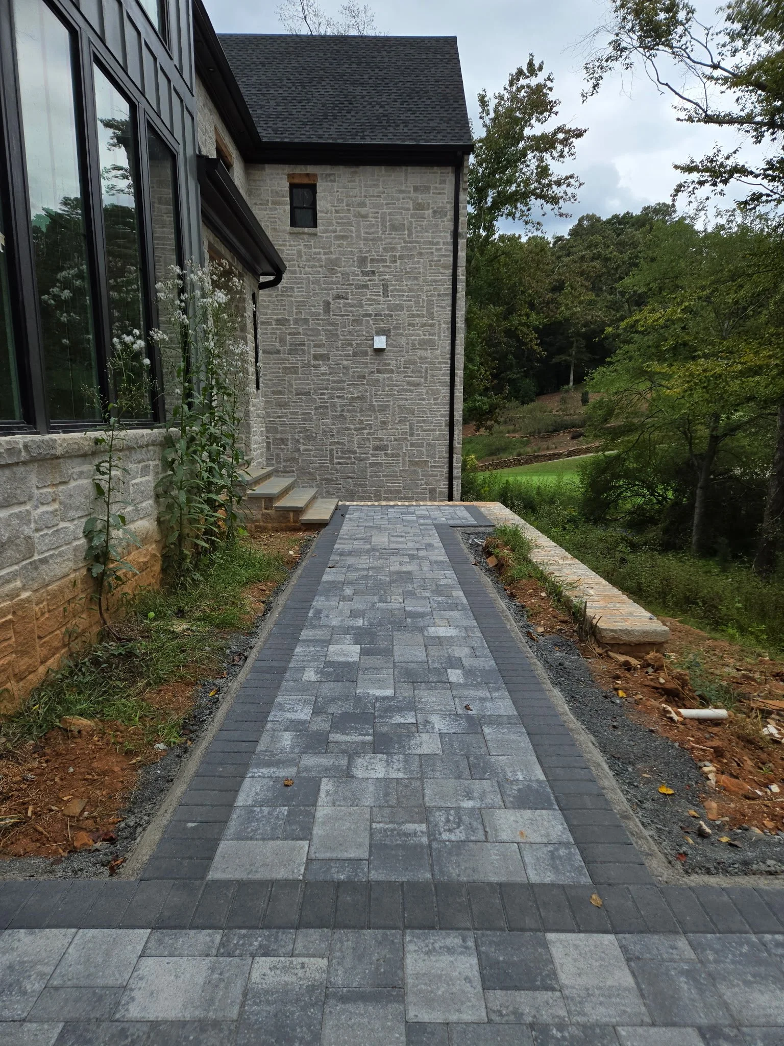 A paved walkway leading to a house with stone walls and large windows, surrounded by grass, trees, and a hillside.
