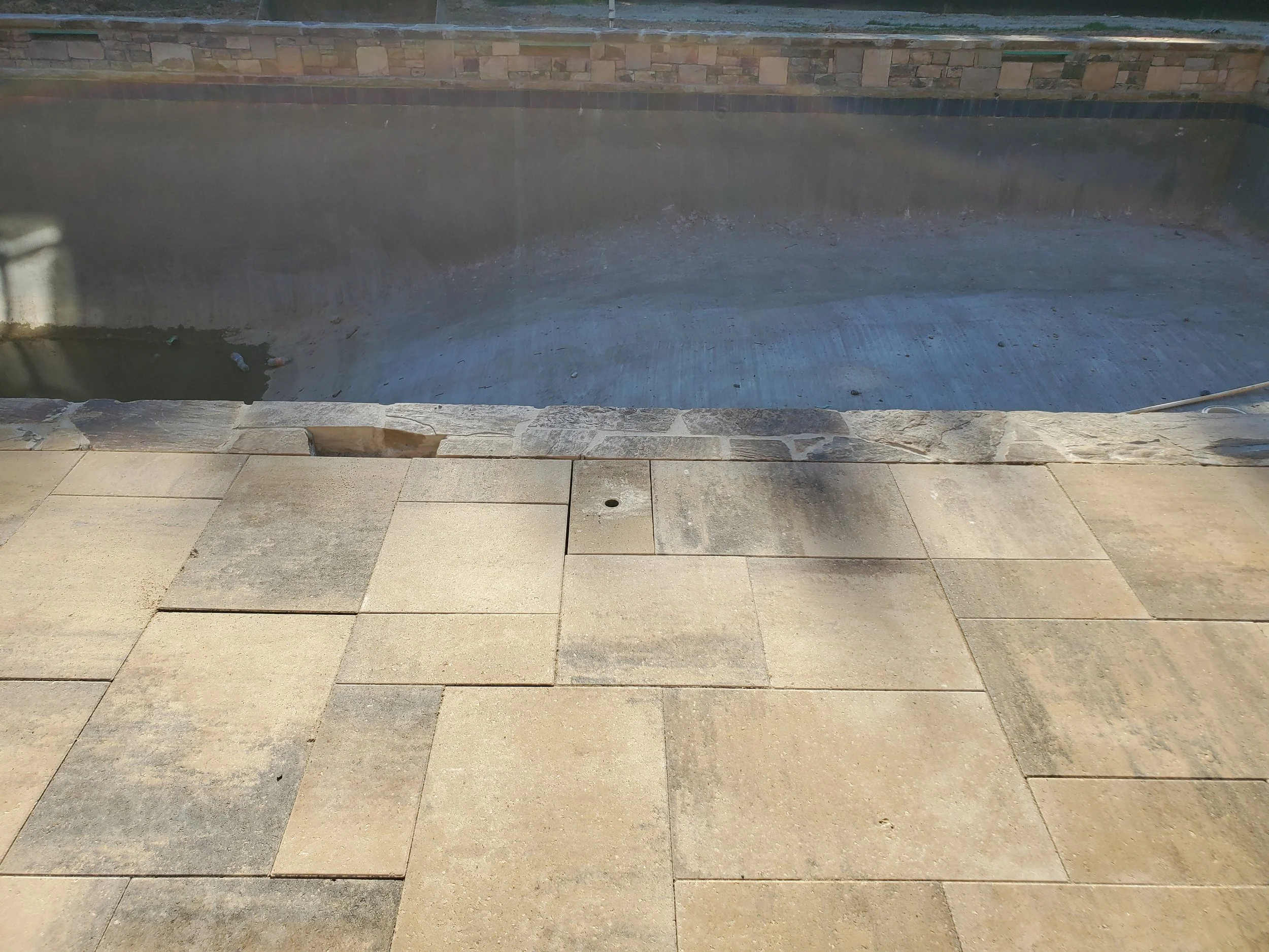 Empty pool with a stone wall at the far side, partially filled with water, with tiles and concrete surrounding it.
