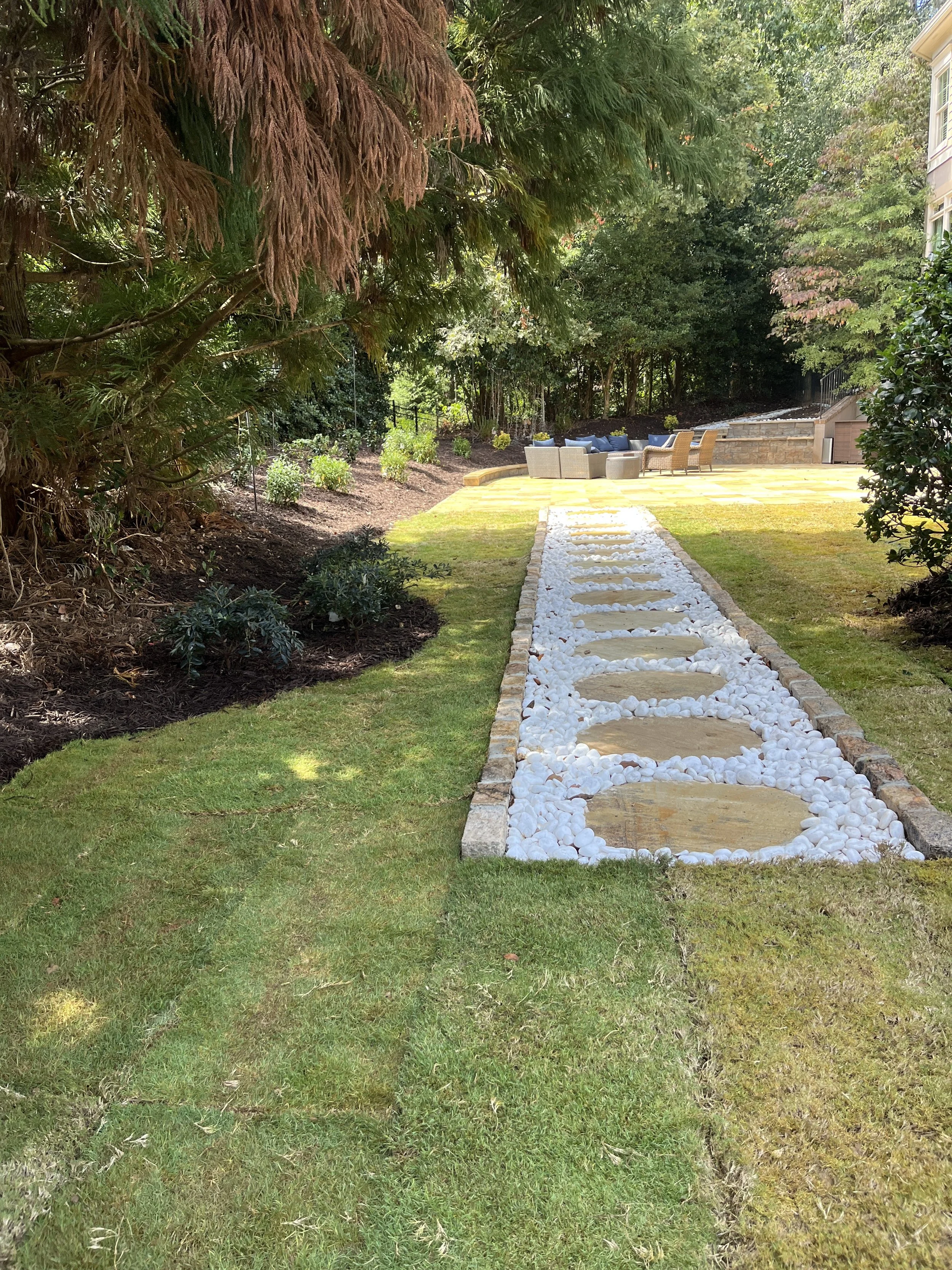 Backyard with a stone pathway surrounded by green grass, trees, and outdoor seating area with chairs and a table in the background.