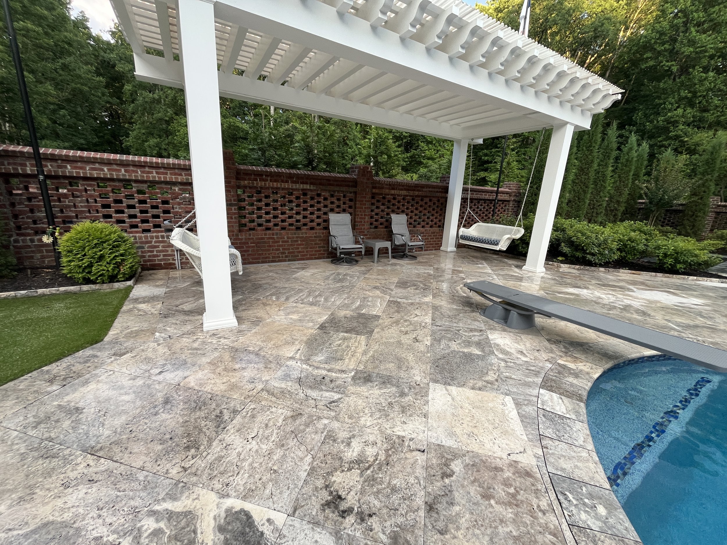 Backyard patio with a pergola, two lounge chairs, a swing, a small table, and a swimming pool with a tiled border, surrounded by greenery and a brick wall.