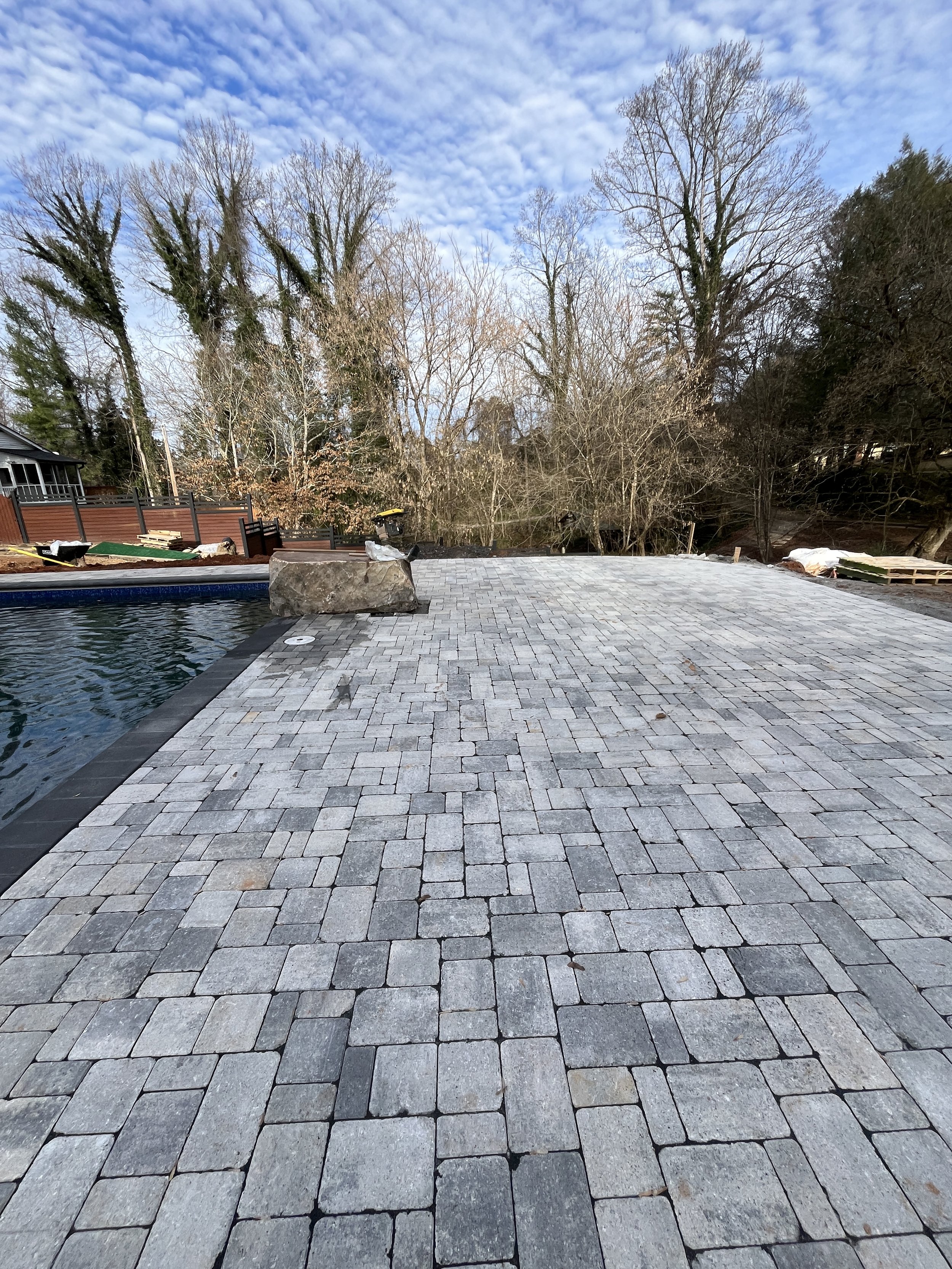 An outdoor backyard scene with a paved stone patio, a swimming pool edge, a wooden fence, leafless trees, and a partly cloudy sky.