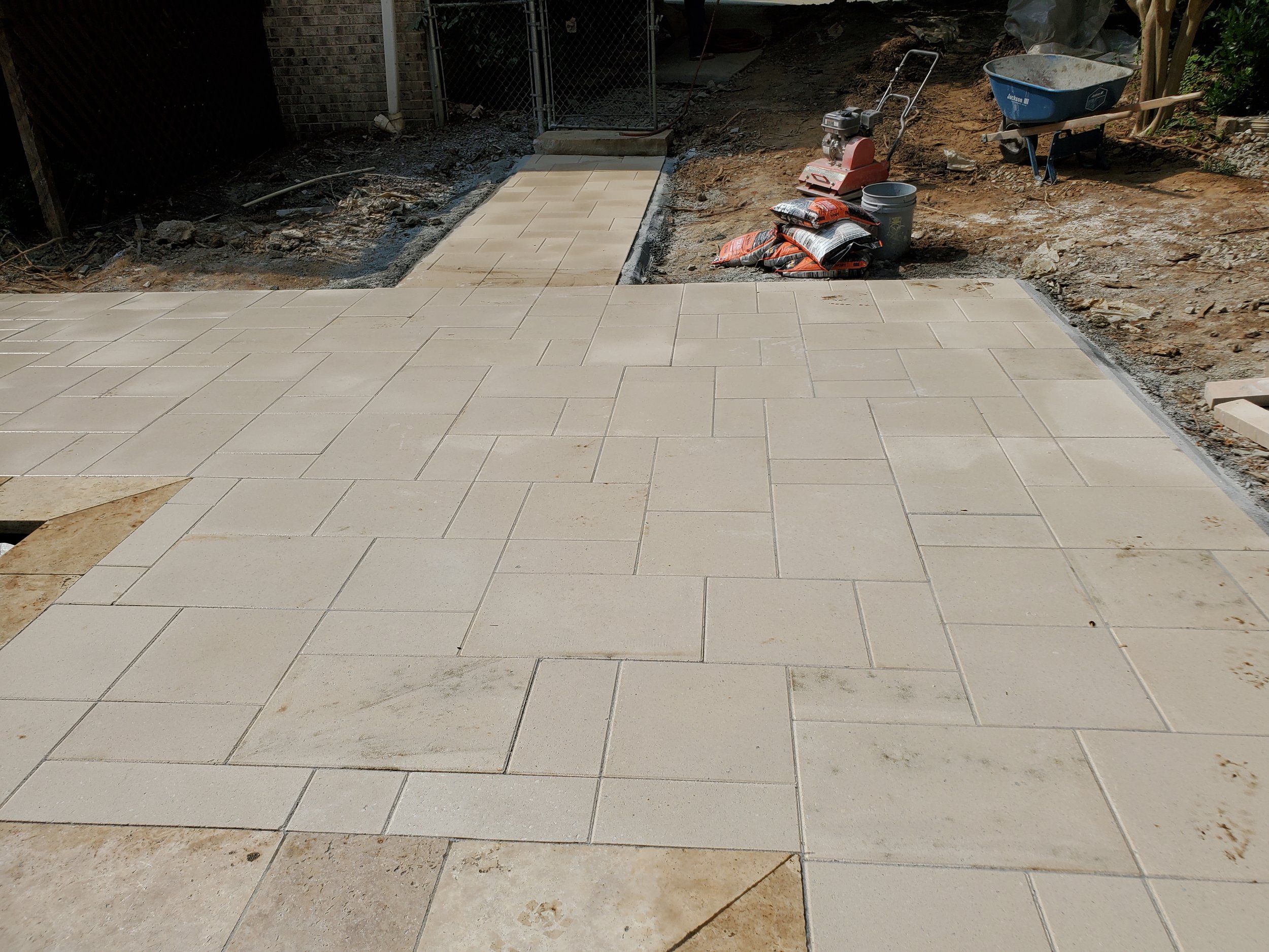 Under construction patio with partially installed light-colored tiles, construction materials, and gardening tools in the background.