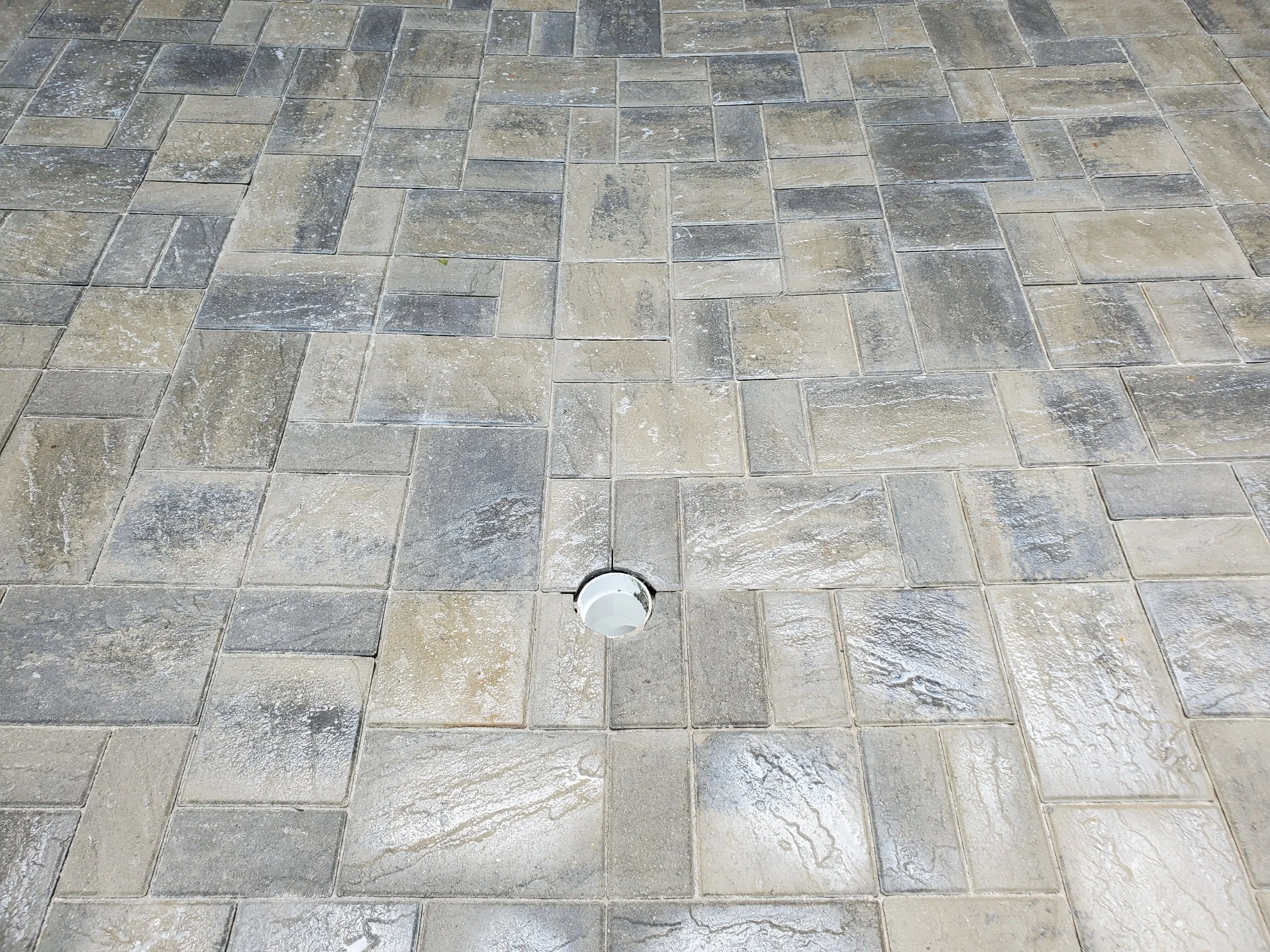 A paved surface with rectangular stone tiles in shades of gray and beige, featuring a drain opening near the bottom center.