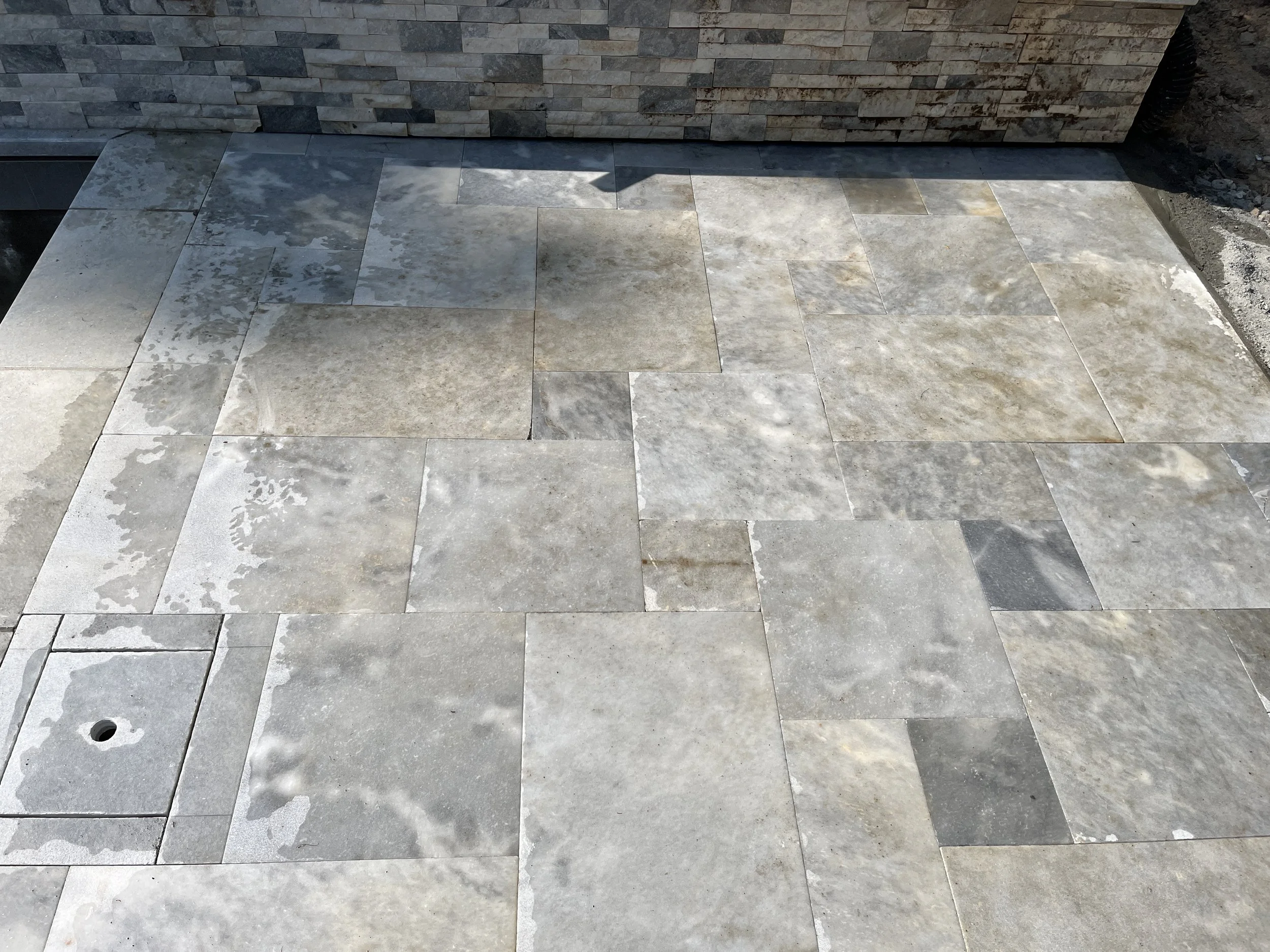 Gray rectangular stone tiles on ground with some water spots and a small hole, next to a brick wall and dirt area on the right.