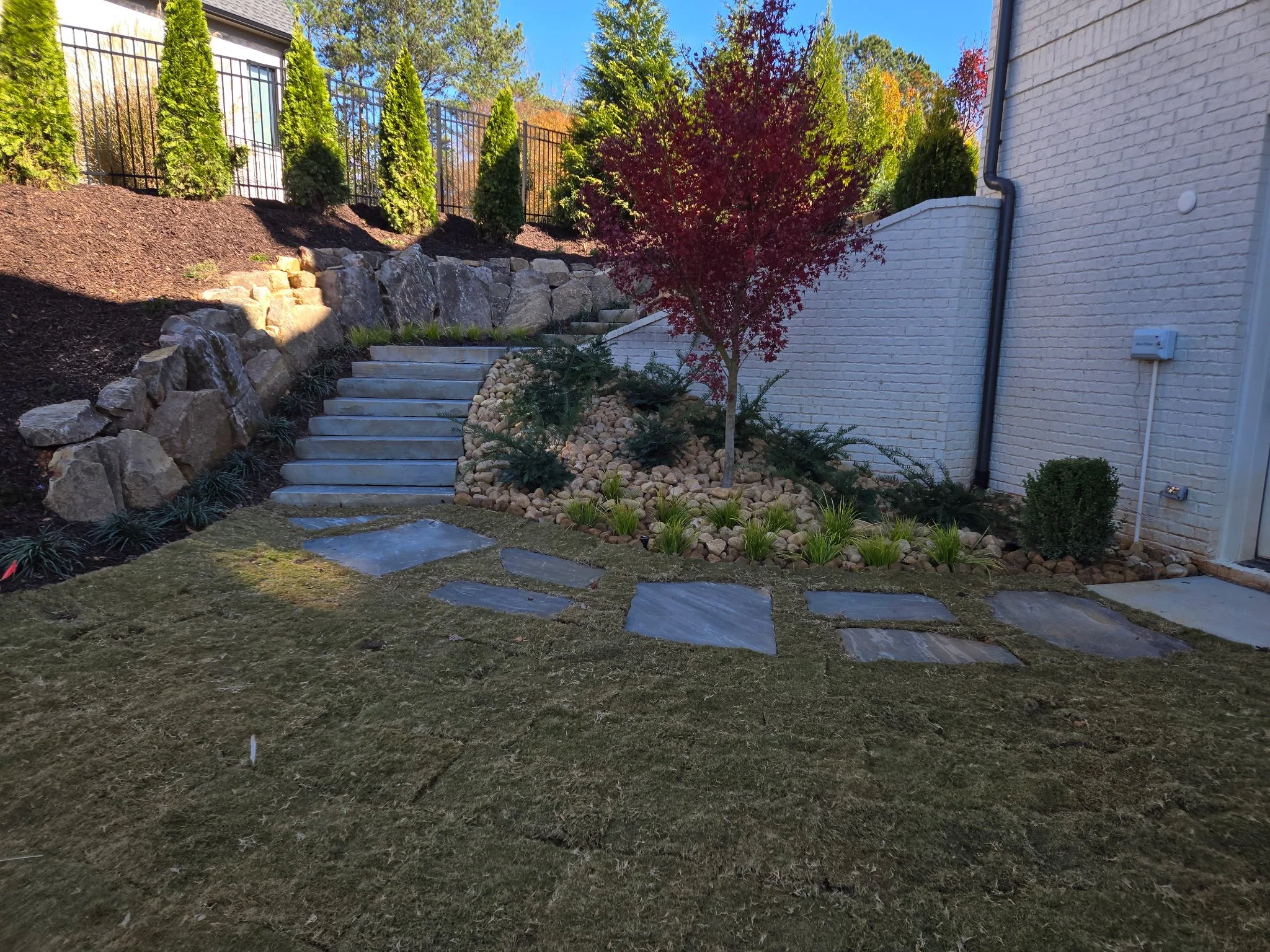 A backyard landscape with stone steps leading up to a garden area with trees, bushes, and a black metal fence. The yard has a brick house wall on the right and a manicured lawn with stone paving in the foreground.
