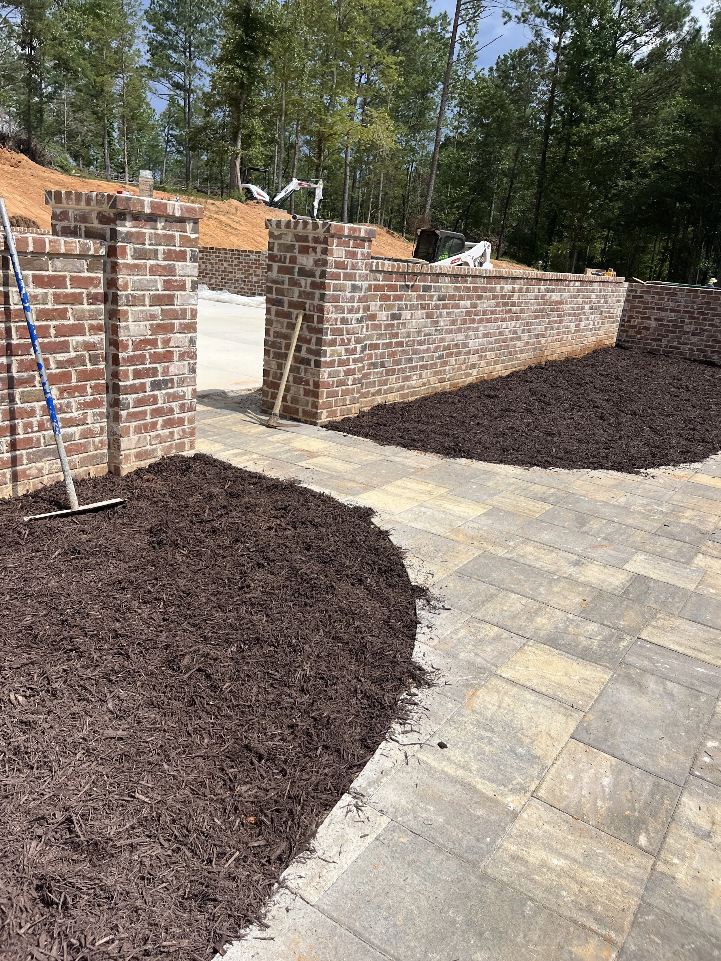 A brick wall under construction with a walkway and mulch in a backyard landscape surrounded by trees.