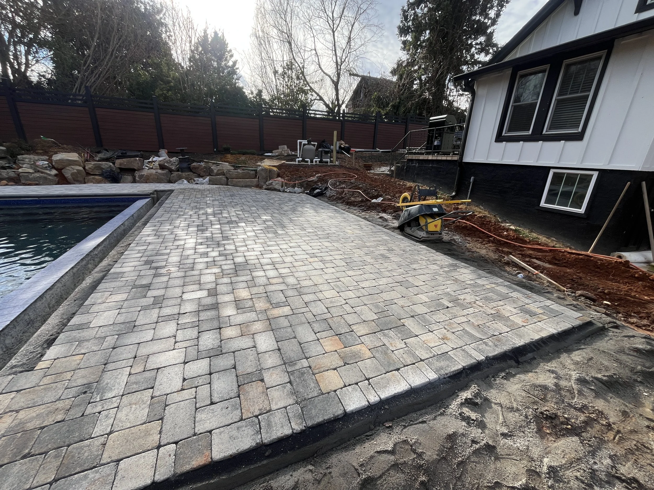 Backyard construction scene showing a paved stone patio, a partially finished garden with rocks and tools, a house with white siding and black window frames, and a brown fence in the background.