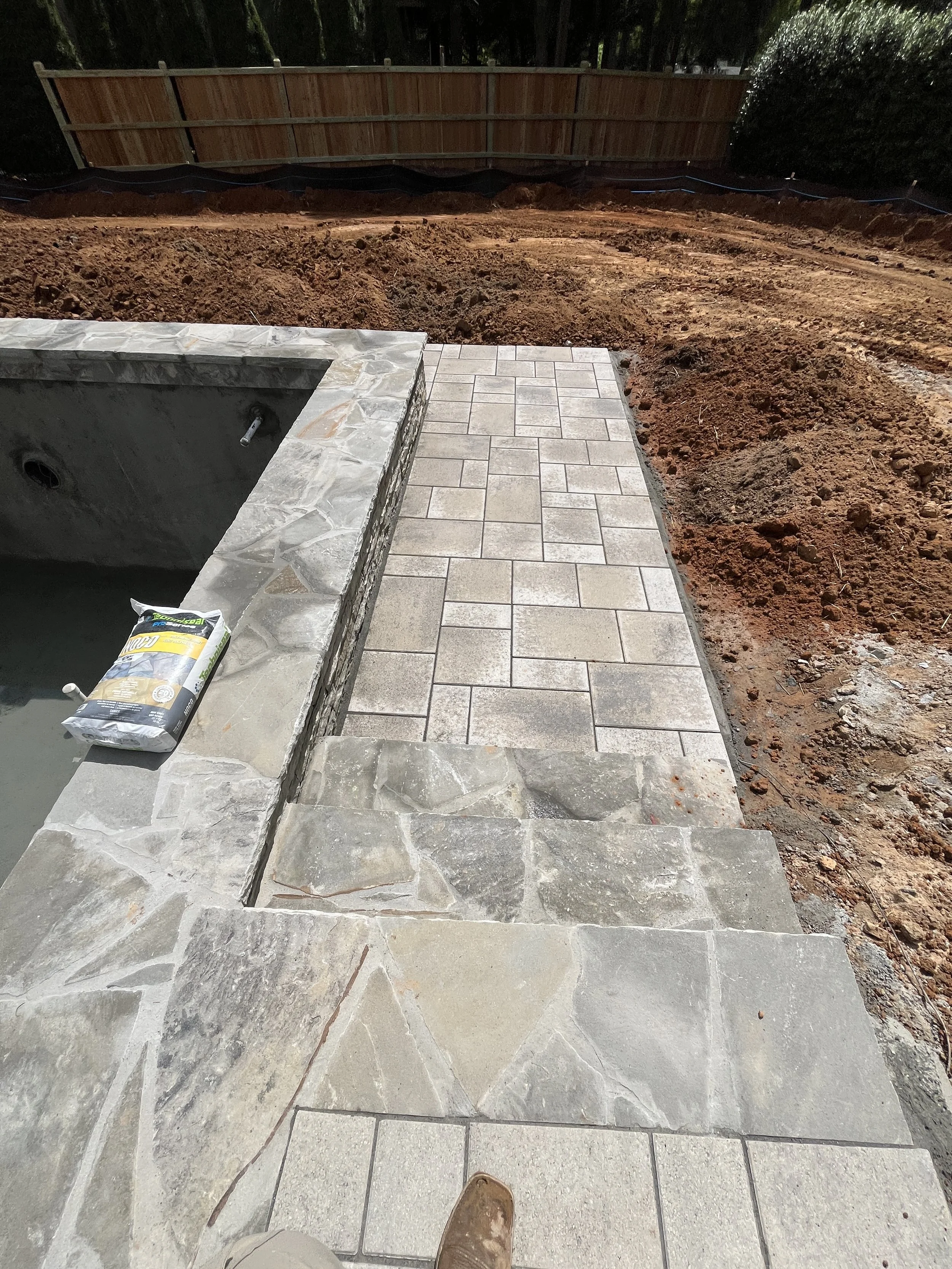 Construction site featuring a partially completed stone patio and foundation with stacked stone border, loose dirt, and a backyard fence in the background.