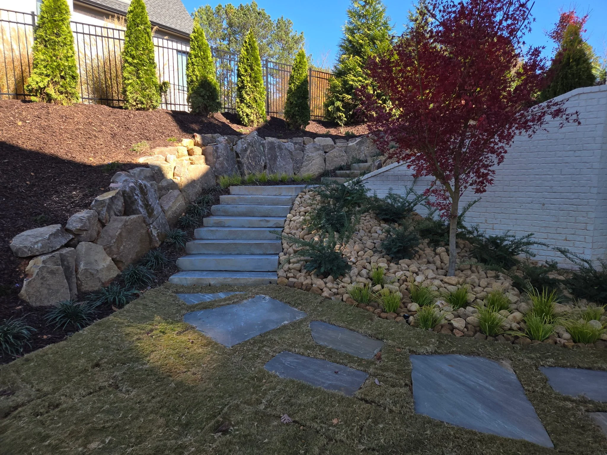 A landscaped backyard with stone stairs leading up a slope, bordered by rocks, with neatly planted bushes and trees, including a small red maple, under a bright blue sky.