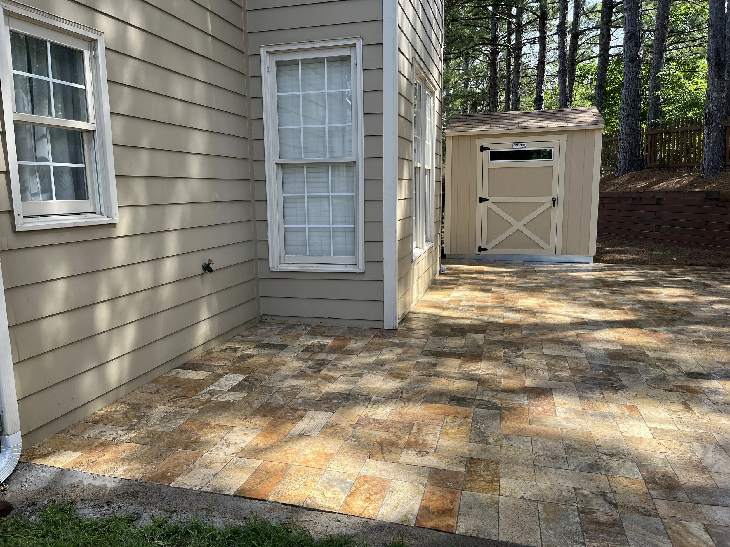An outdoor patio area with multicolored stone tiles next to the beige siding of a house, with a window and a water spigot visible, and a small storage shed in the background surrounded by tall trees.