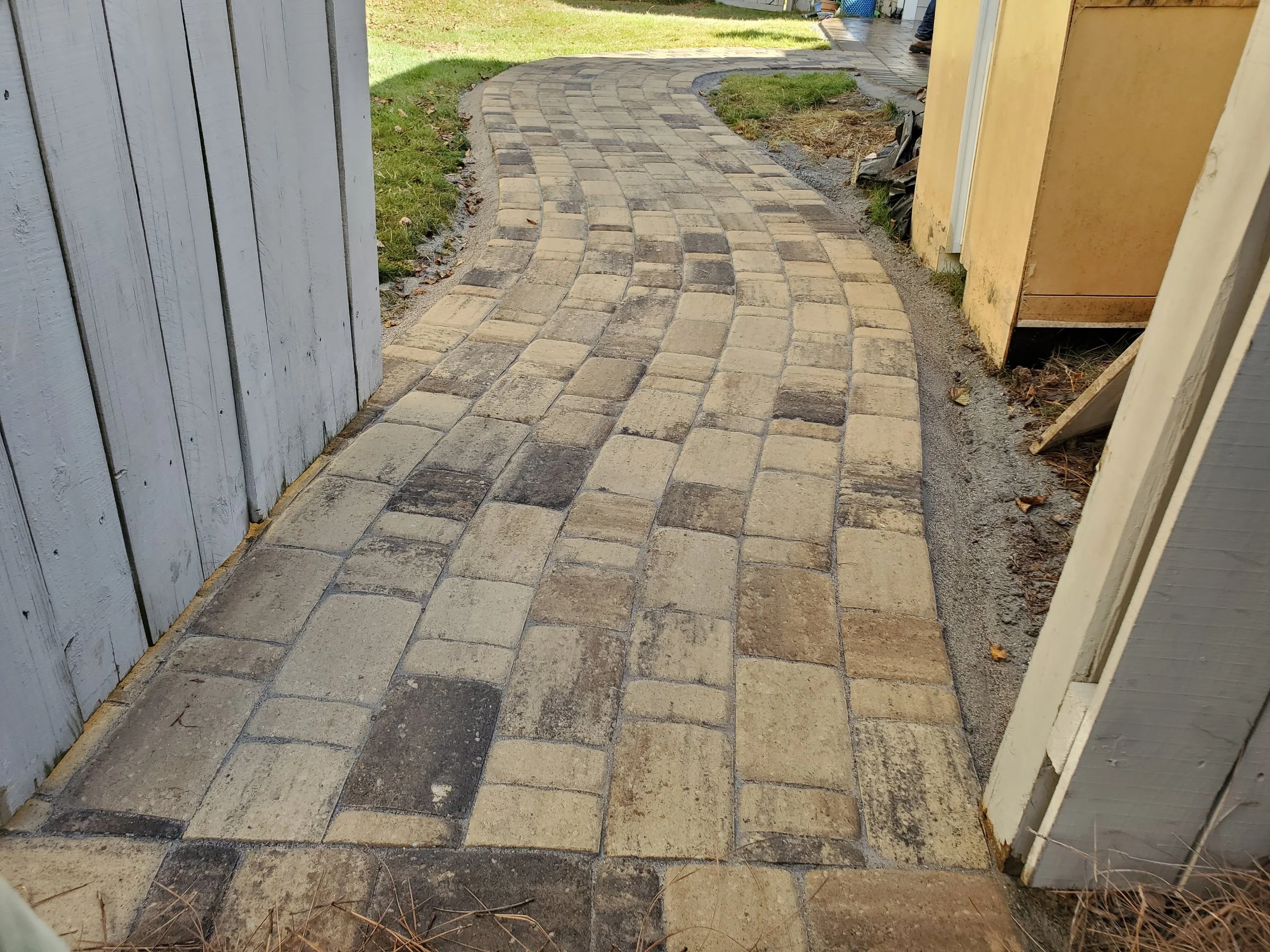 A newly paved brick pathway curves from a white wooden fence on the left to a yellow house on the right. The pathway is bordered by grass and soil, with a person visible in the background near a garden hose and tools.