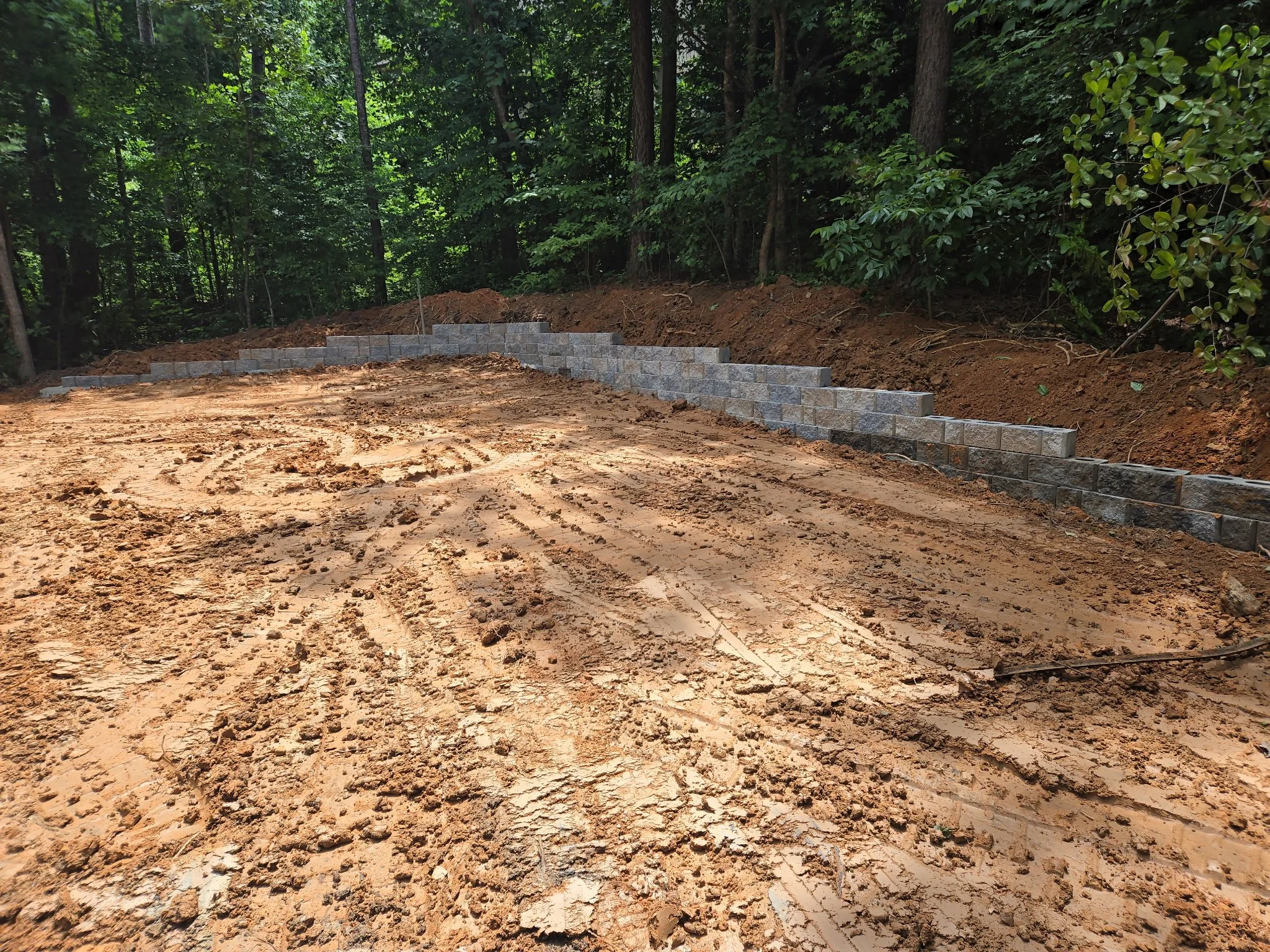 A dirt pathway with freshly laid concrete blocks forming a small stone retaining wall on the right side, surrounded by green trees in a forest setting.